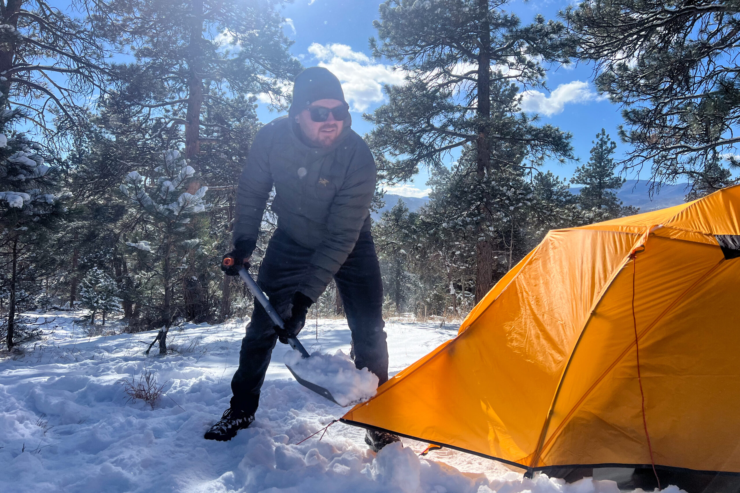 Person wearing the Arc'teryx Cerium Hoody while shoveling snow near an orange tent in a snowy forest setting.