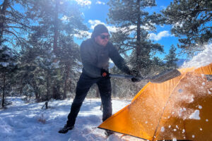 A man shoveling snow from around the vestibule of his tent. It's a sunny day, and snow is flying through the air from the shovel.