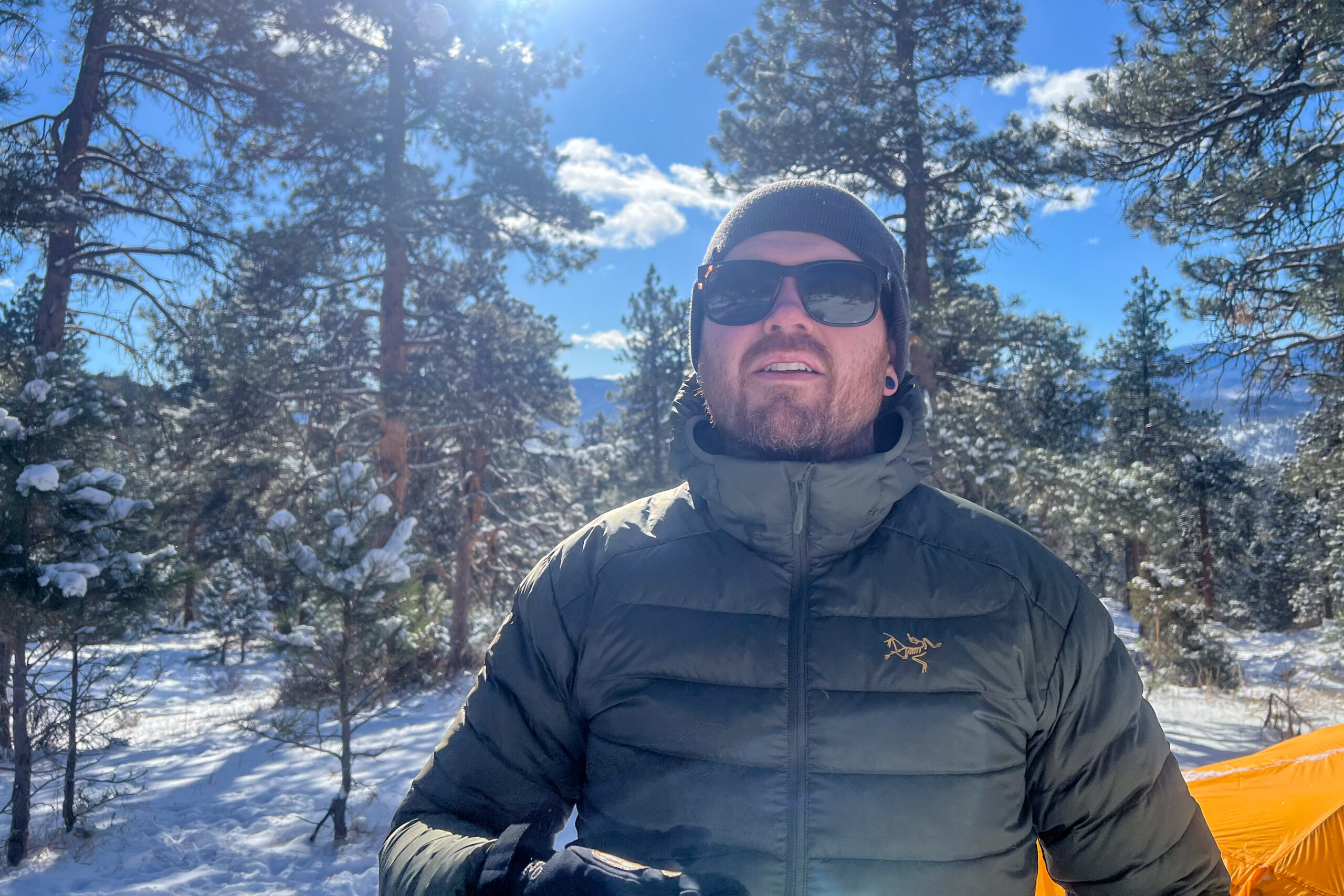 A man stands in a snowy forest with the sun in the sky behind him. He's facing the camera and wearing a green down jacket.