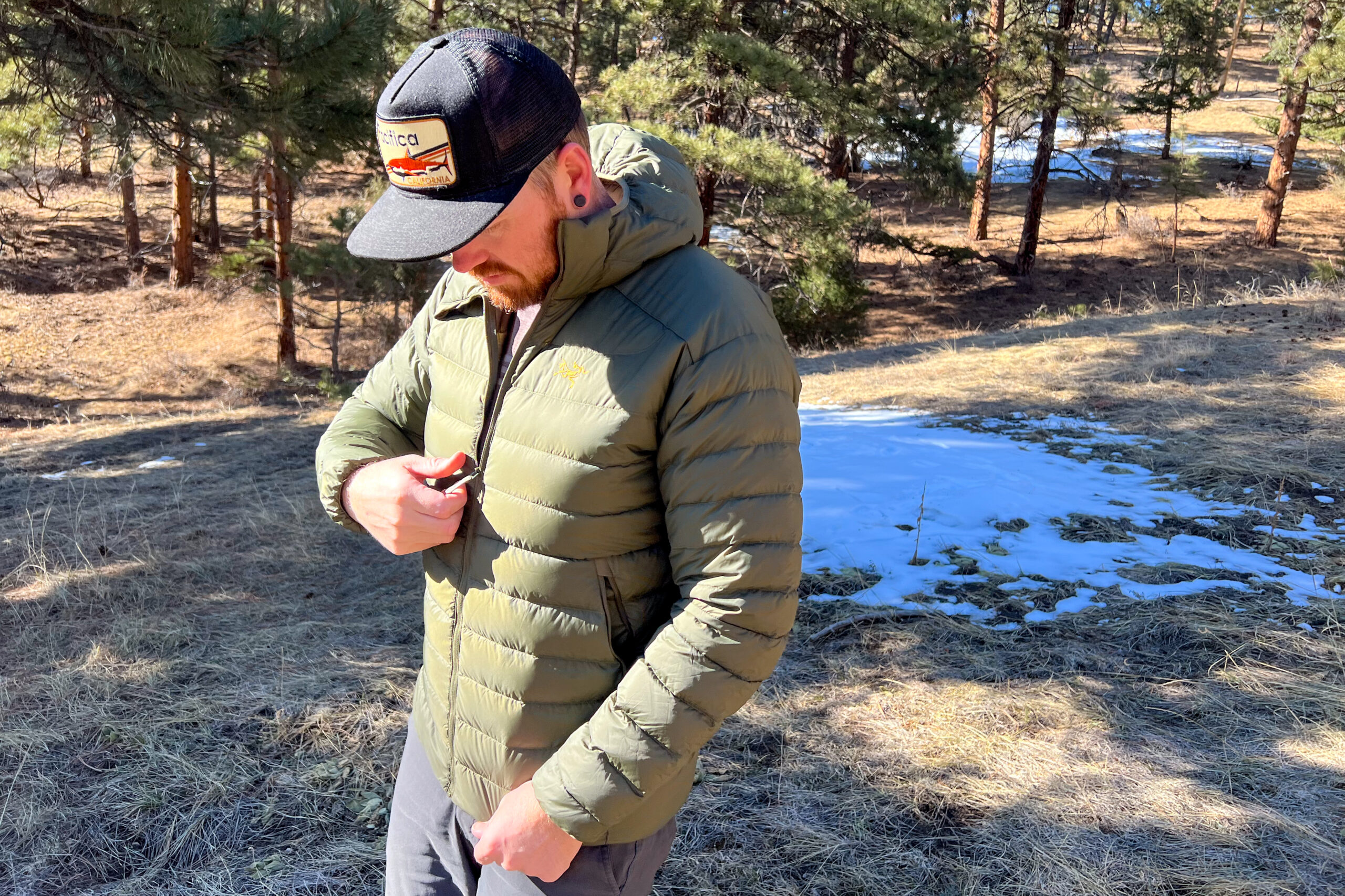 A man walking in the forest. He's zipping his jacket and looking down. There are small snow patches in the background.