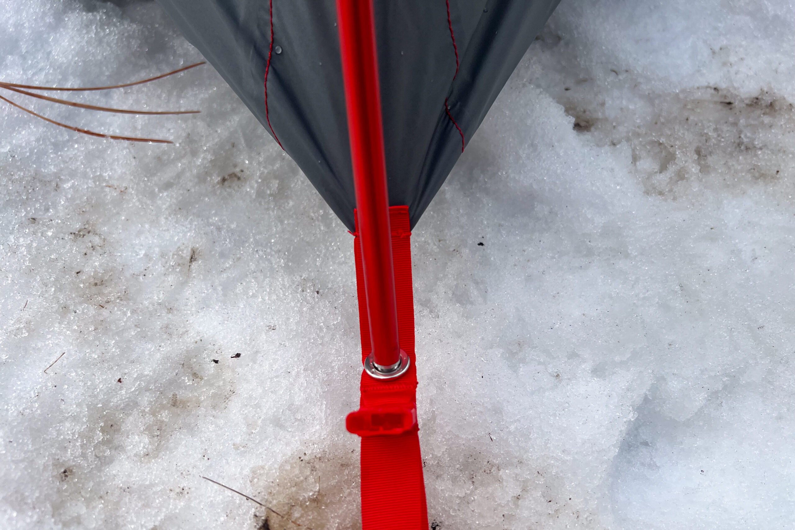 A detailed view of a red tent pole secured to the tent's corner grommet and webbing strap in snow.