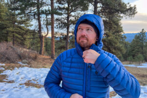 A person adjusting the zipper on the Black Diamond Approach Down Hoody, with the hood up, standing outdoors in a snowy forest setting.