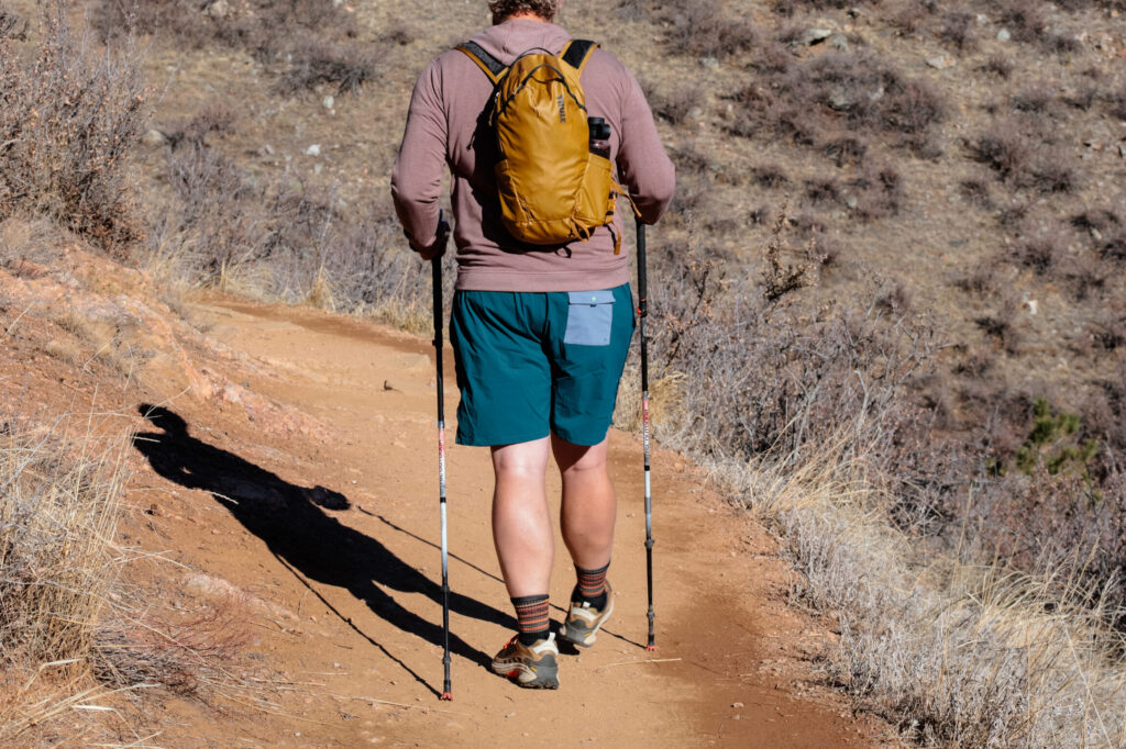 Man hiking down a trail with a pack and trekking poles in green shorts.