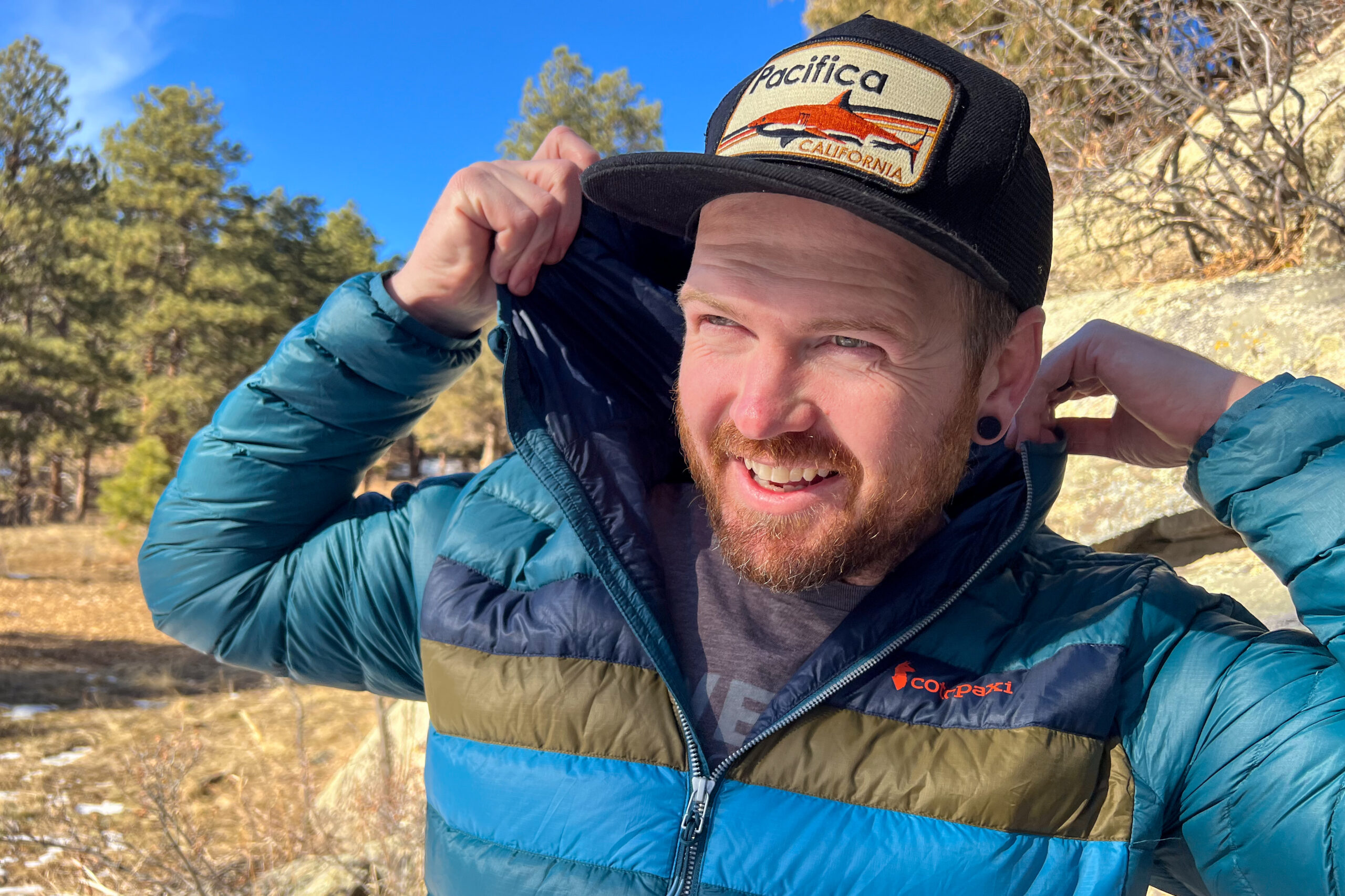 A man in the Cotopaxi Fuego jacket pulls up the hood, smiling and looking off-camera, with trees and rocks in the background.