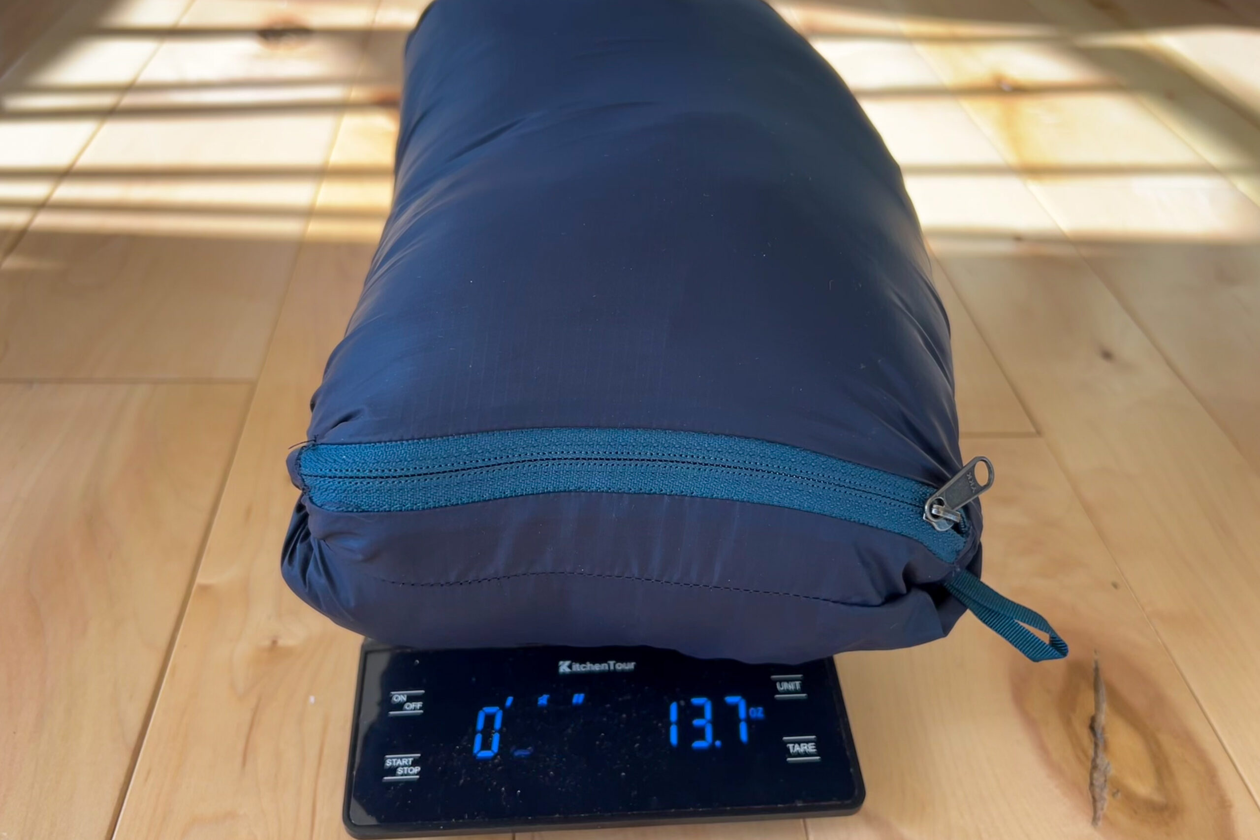 A Cotopaxi Fuego jacket compressed into its stuff pocket sits on a scale, displaying a weight of 13.7 ounces.