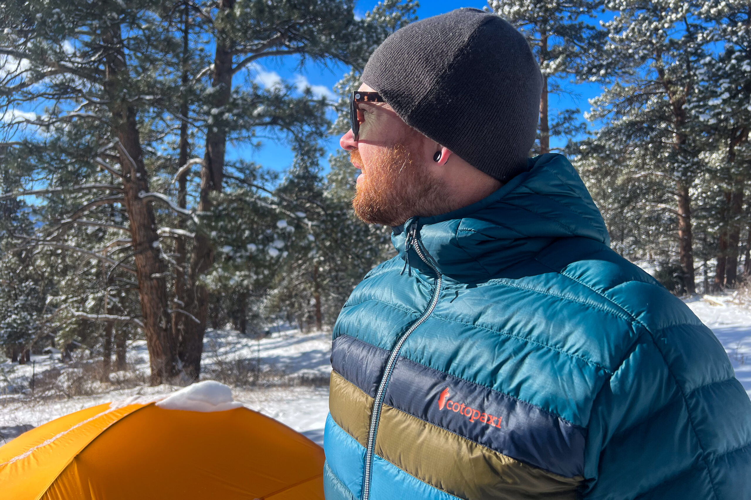 Side profile of a man in a blue Cotopaxi Fuego jacket, looking into the distance with a snowy forest and yellow tent behind him.