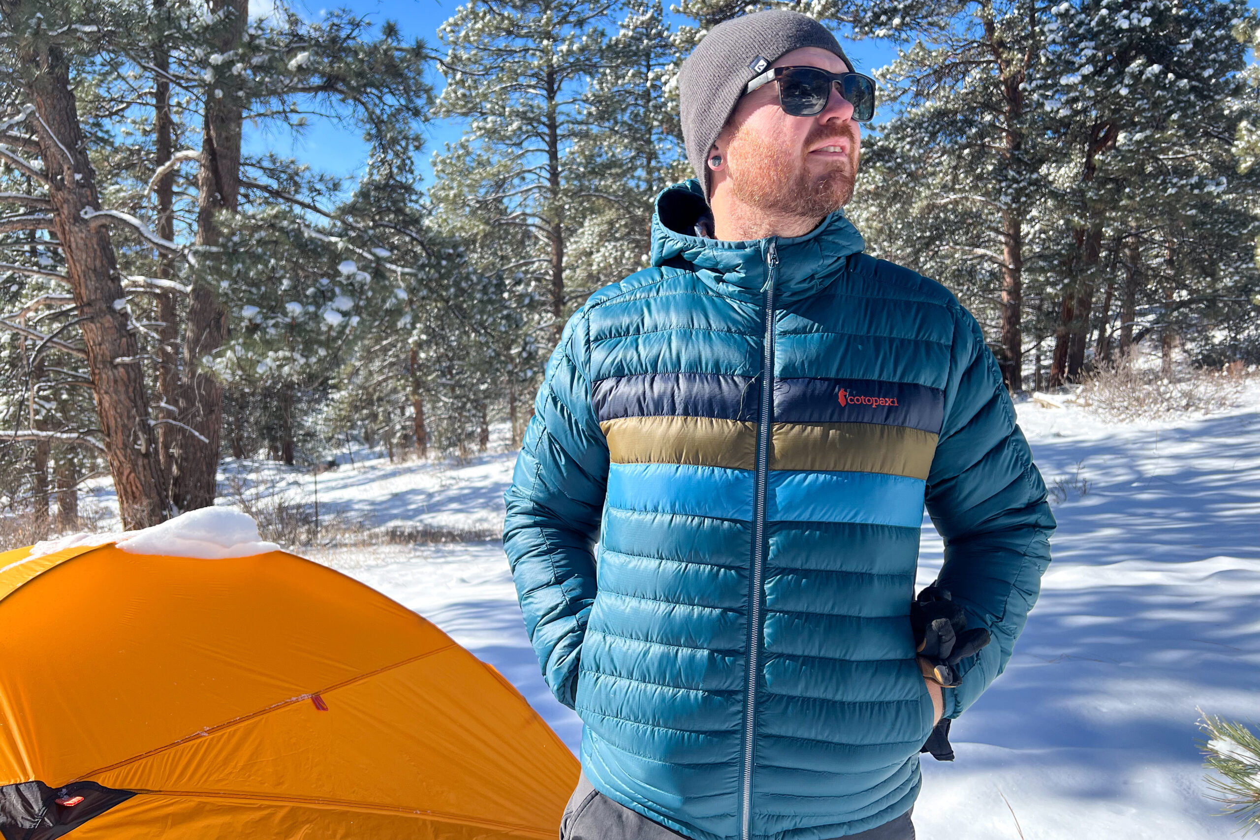 A man in the Cotopaxi Fuego jacket stands confidently in a snowy forest with an orange tent in the background, hands tucked into his jacket pockets.