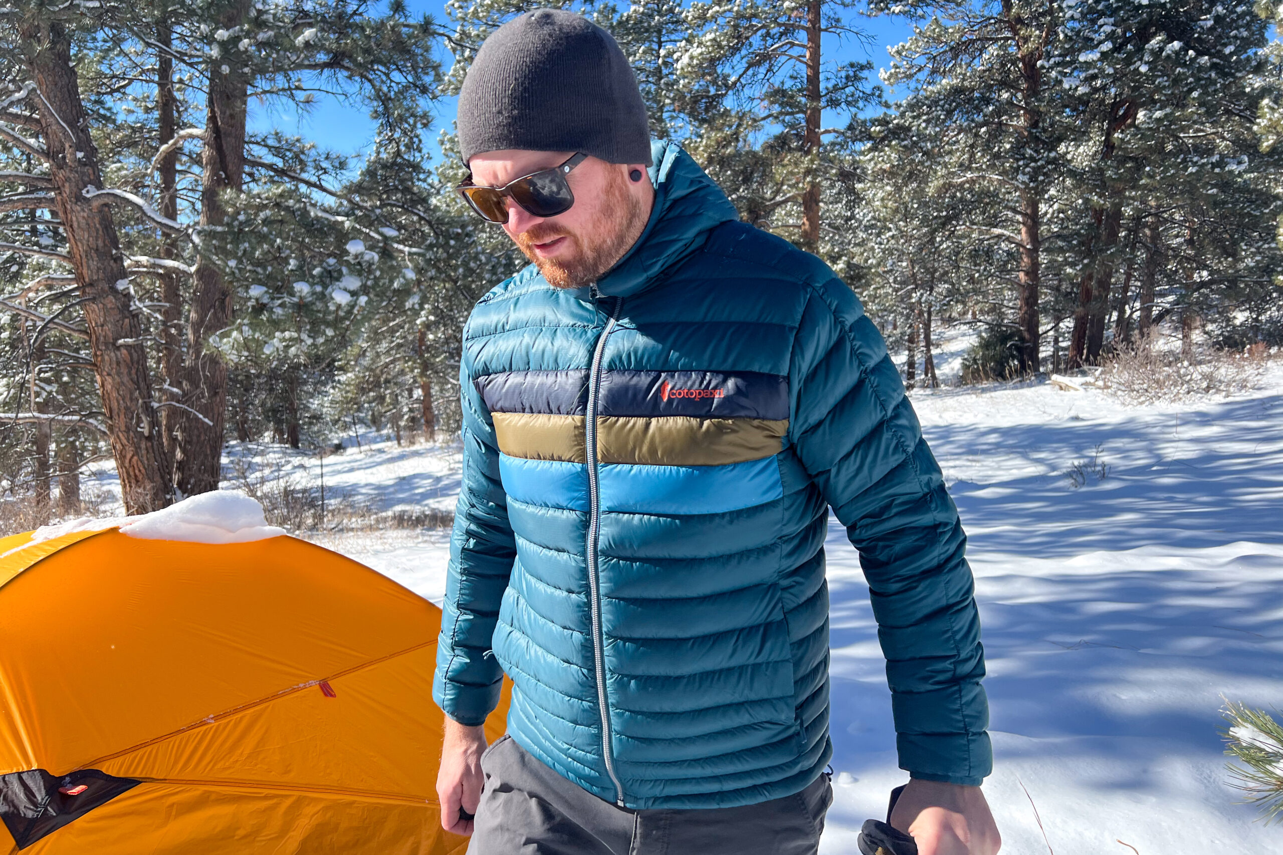 A man in a Cotopaxi Fuego jacket, wearing a beanie and sunglasses, walks past a snow-covered tent in a pine forest.