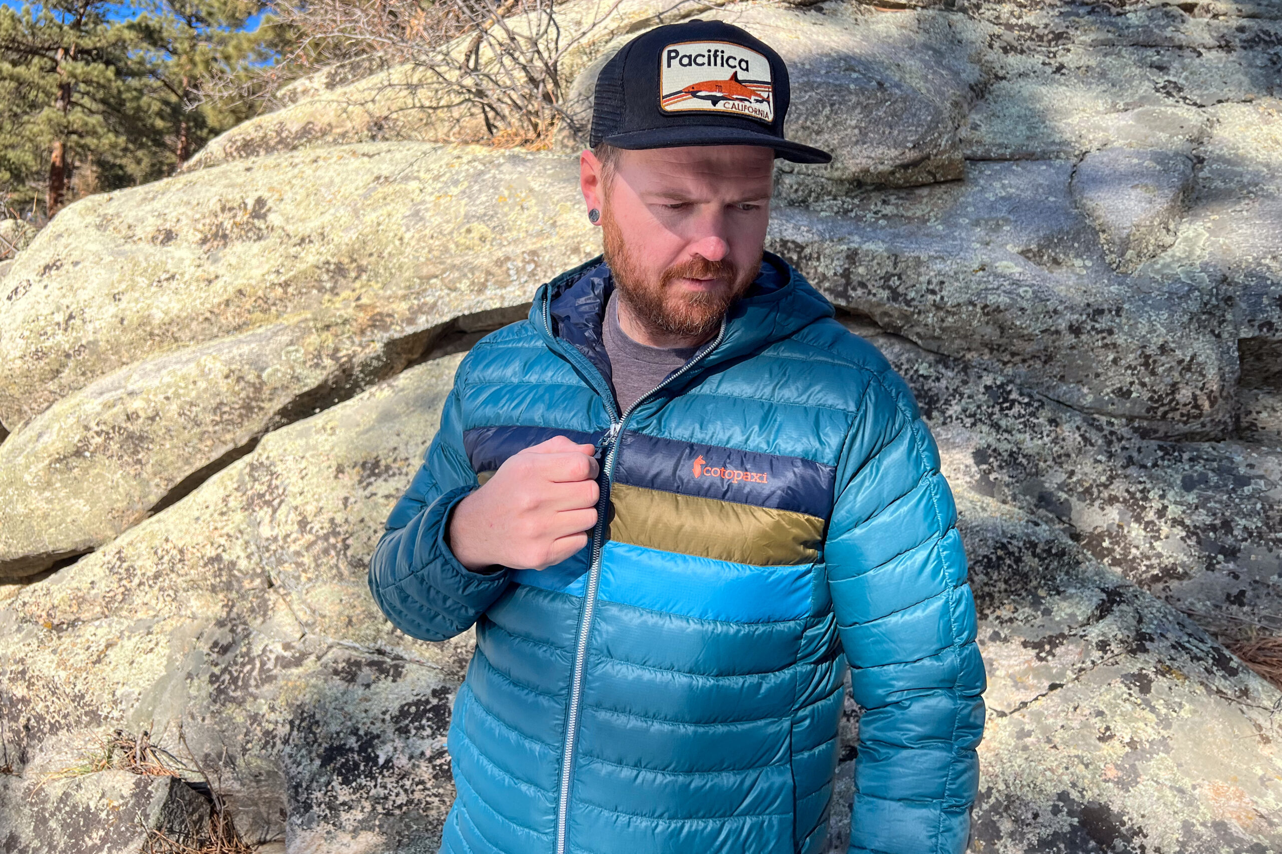 A man adjusts the front zipper of his Cotopaxi Fuego Hooded Down Jacket while standing near textured rock formations outdoors.