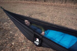 a man in a blue sleeping bag lays in a black hammock