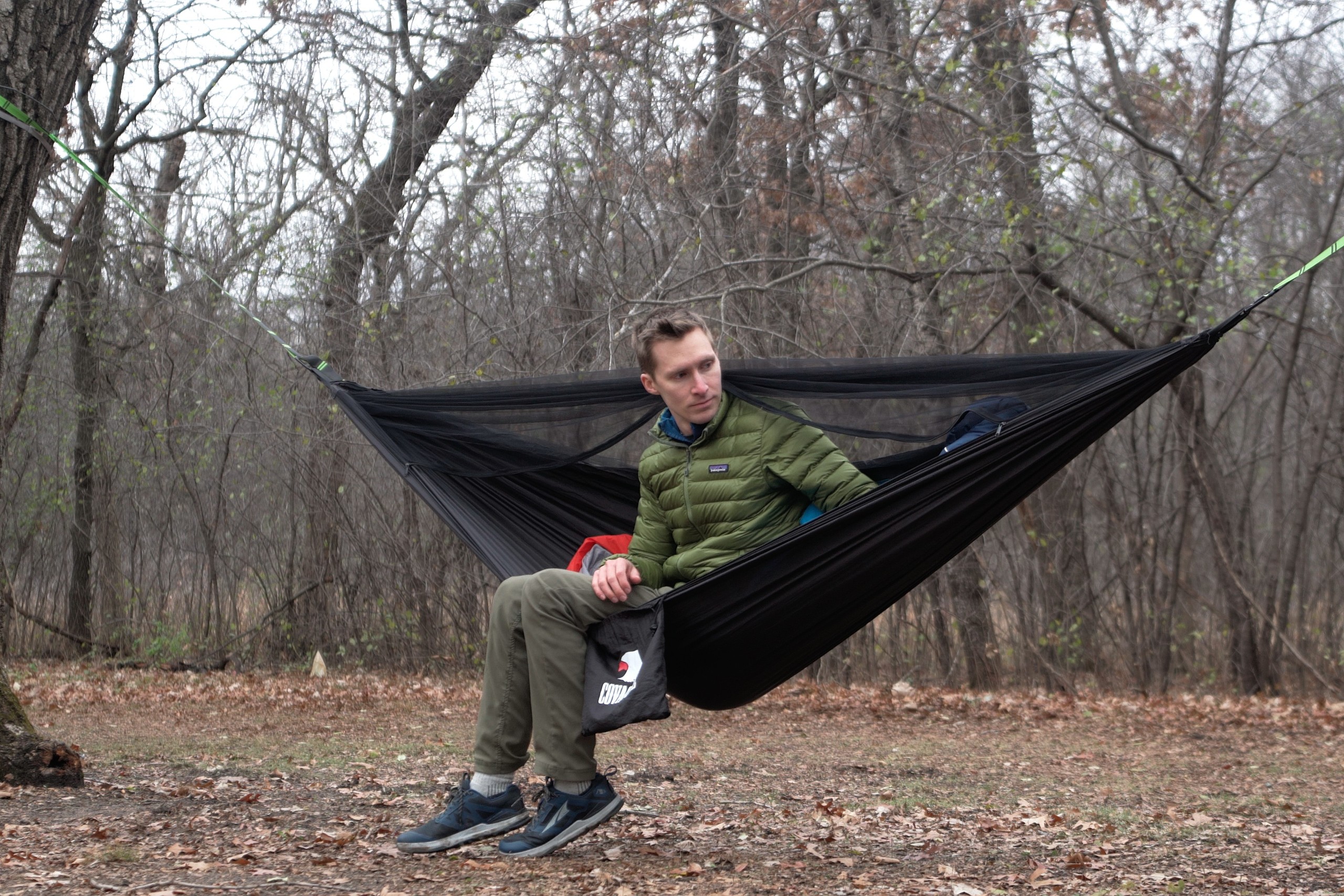 a man in a green coat sits in a black hammock in a clearing in the forest