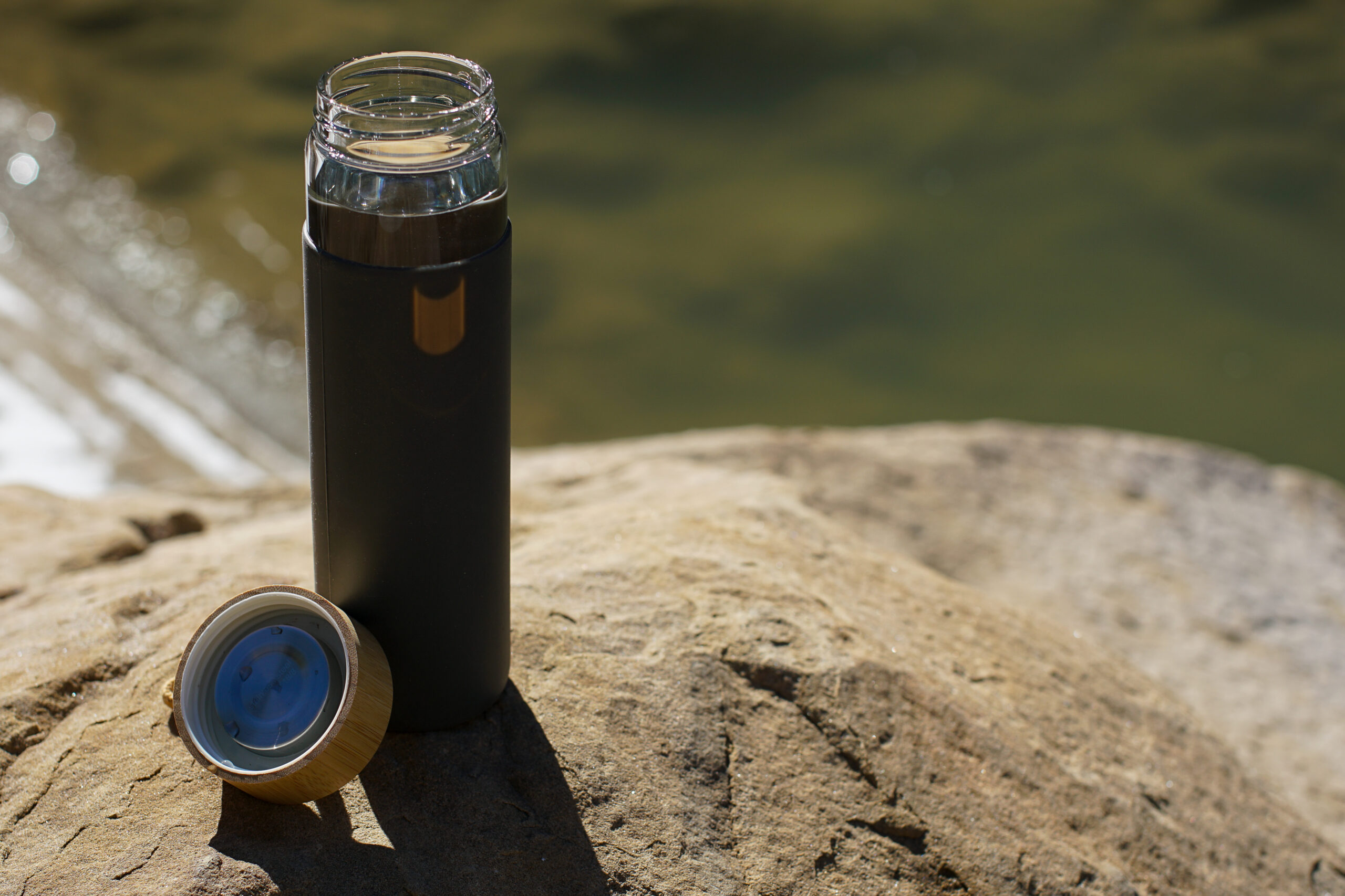 The Yomious Glass water bottle with its bamboo lid beside it, sits on a rock with a river in the background.