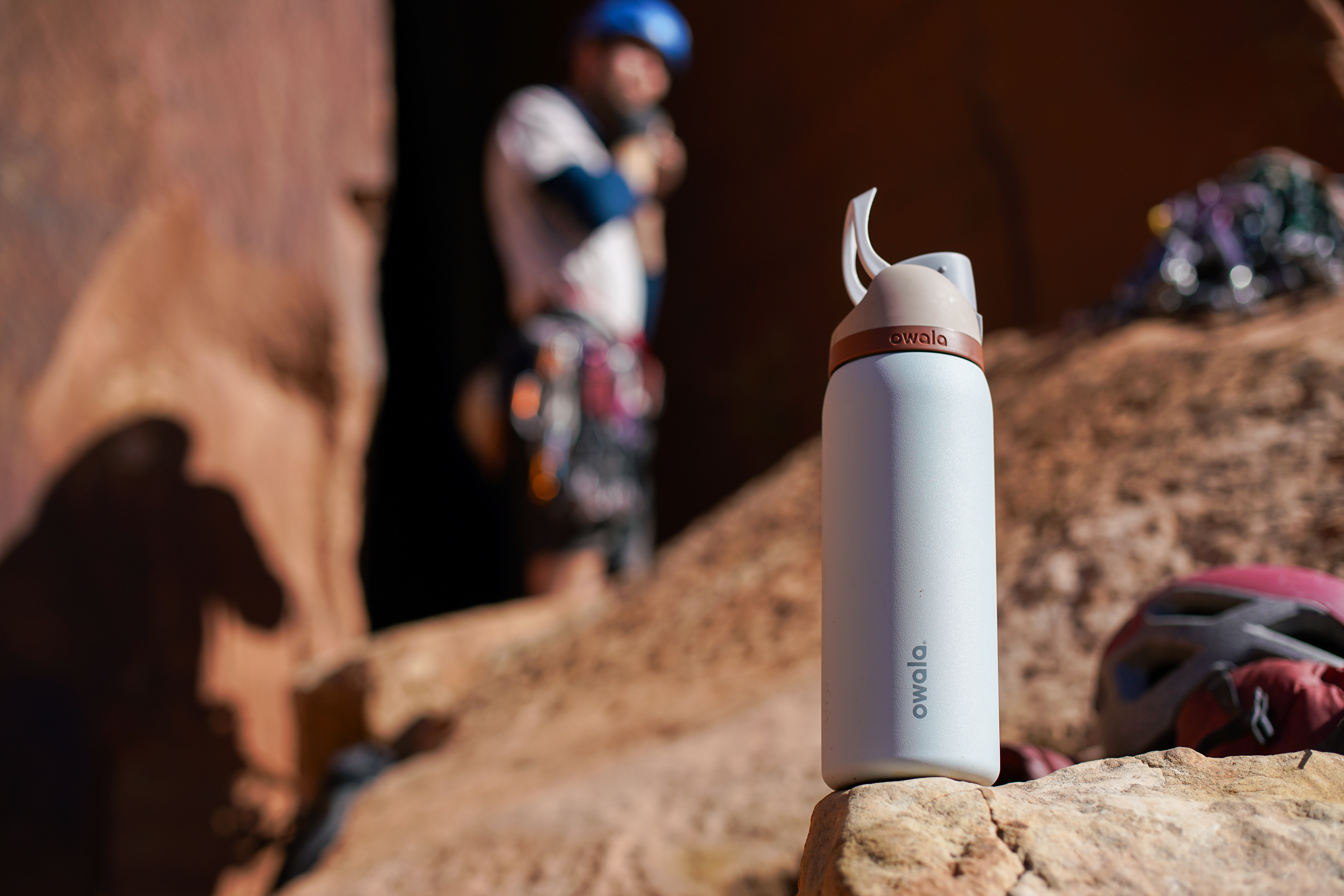 The Owala FreeSip Vacuum Insulated water bottle sits on a rock with a climber in the background.