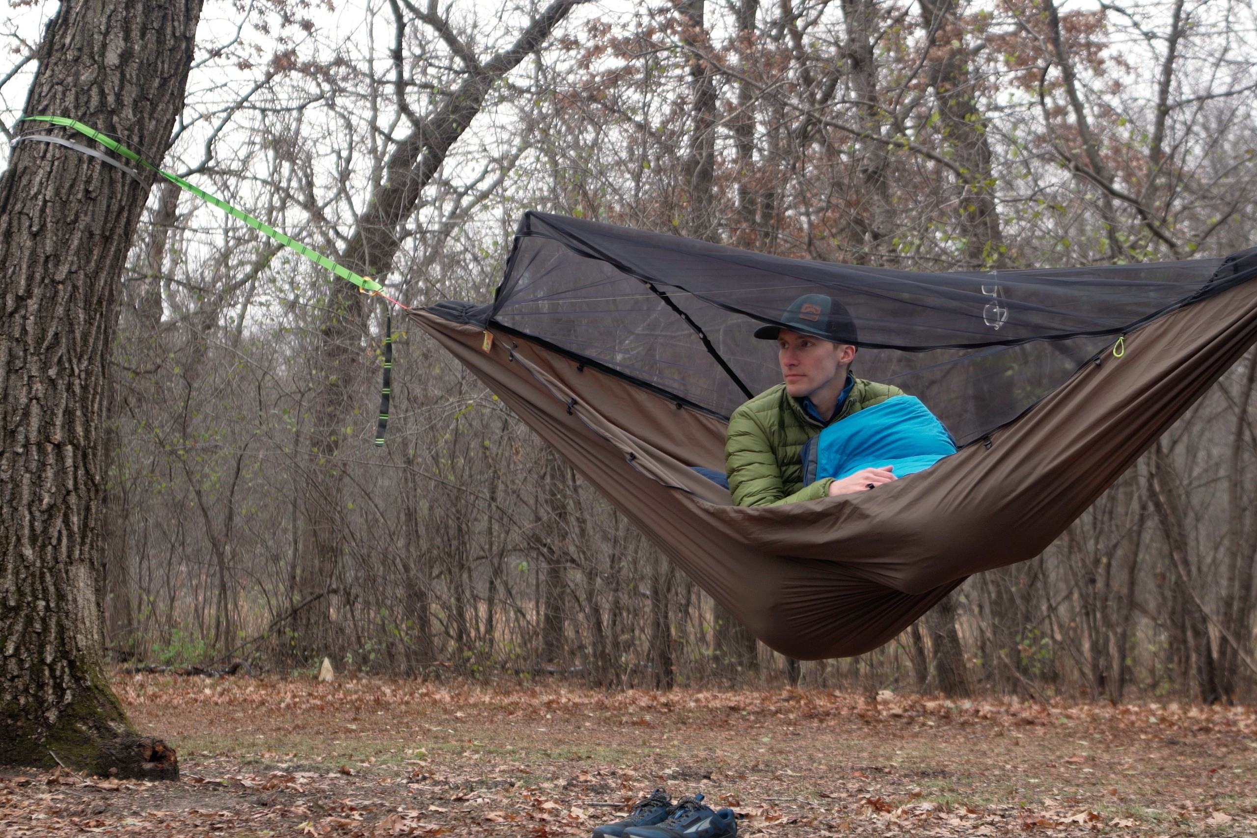 a man in a blue sleeping bag sits up in a green hammock