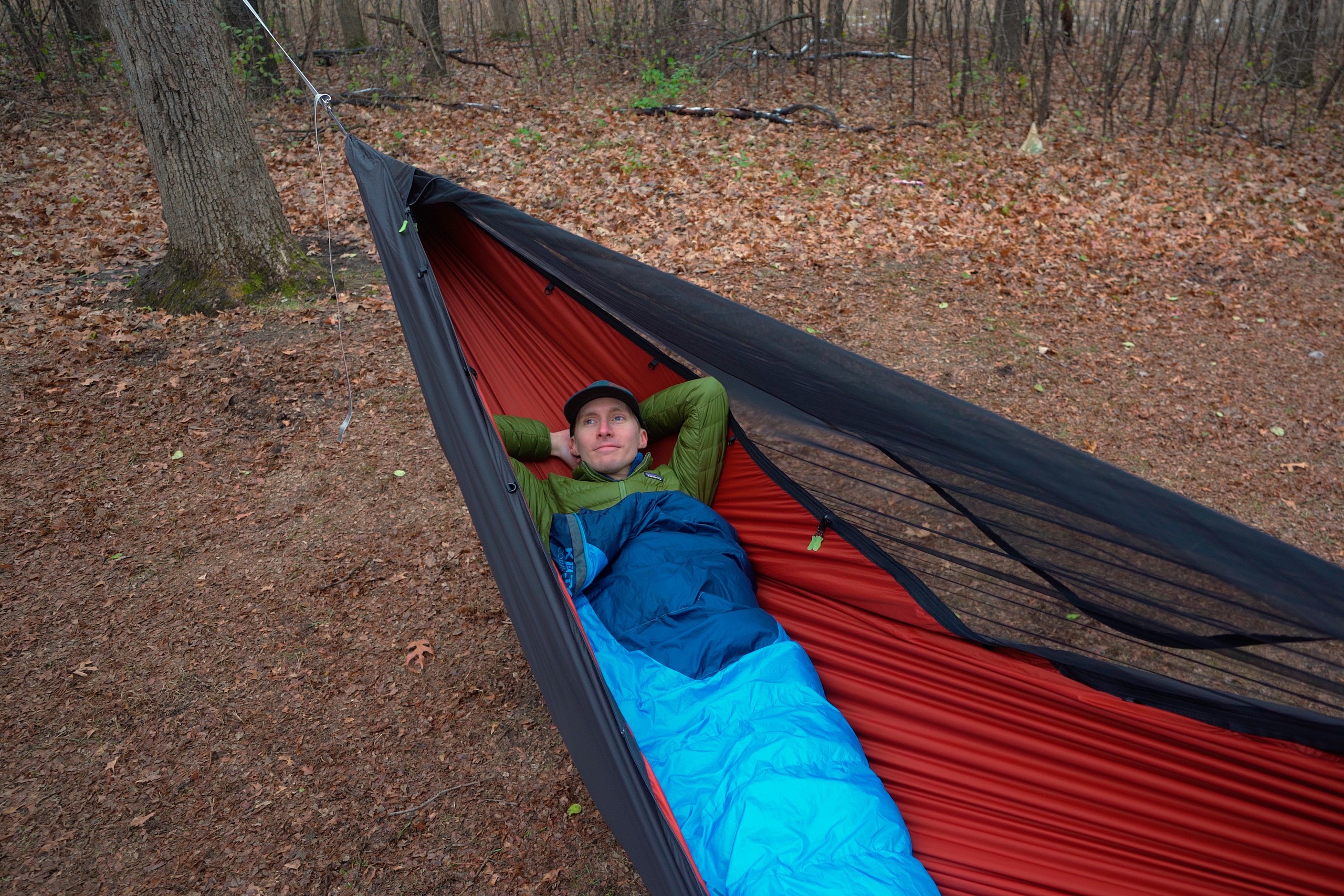 a man in a blue sleeping bag lays back in a red and black hammock