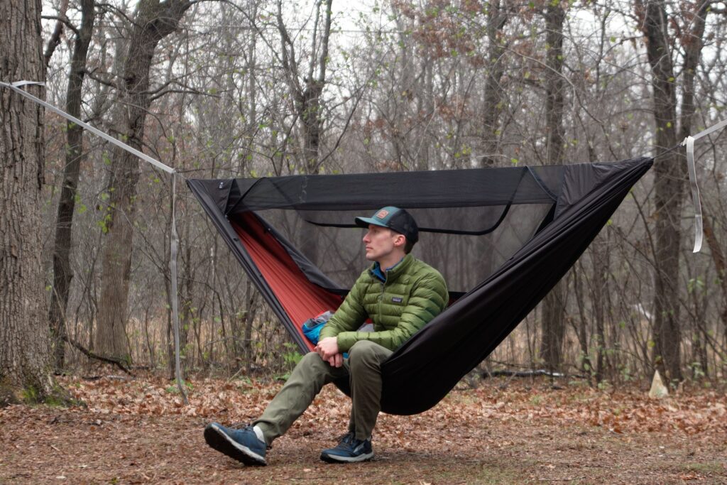 a man sits in a hammock with a red interior and black exterior in a forest clearing