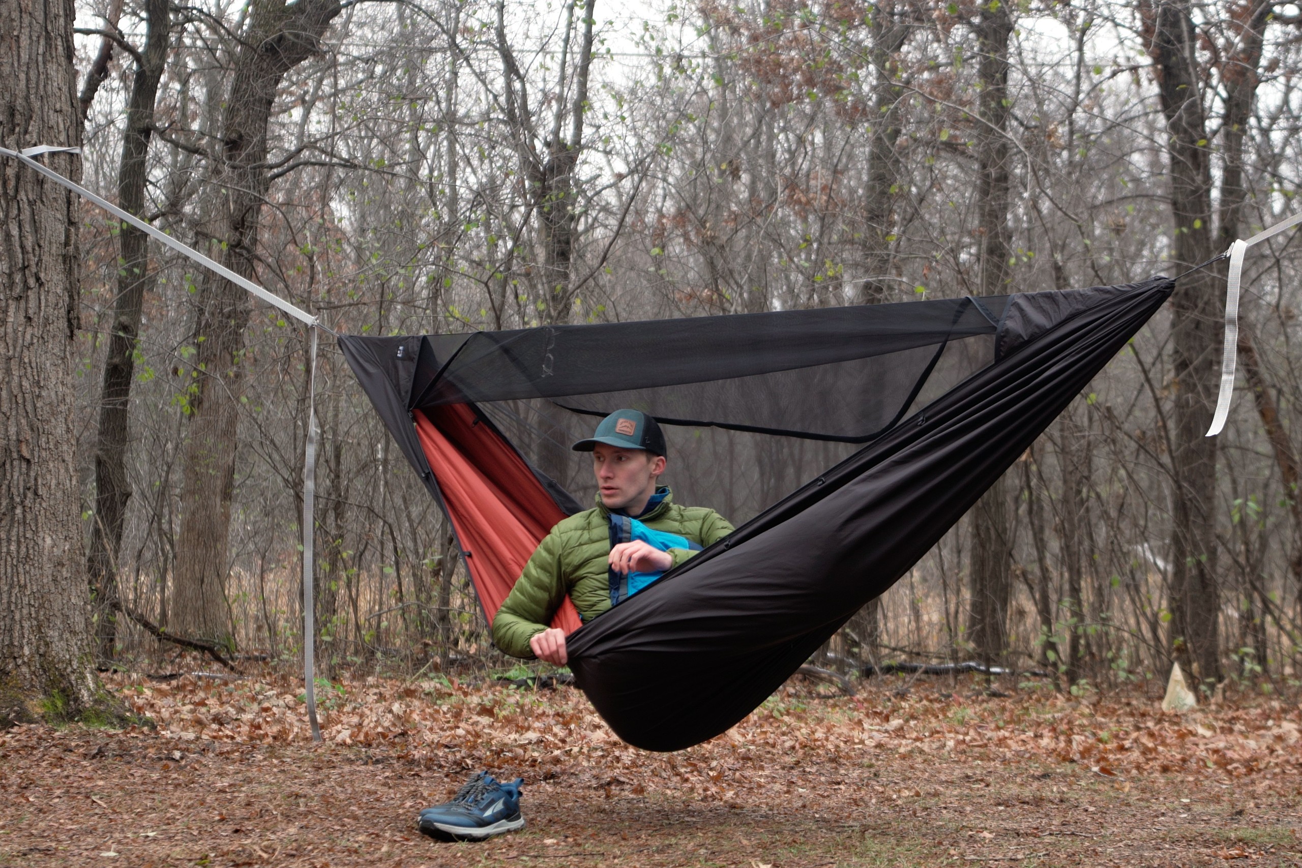 a man sits in a hammock with a red interior and black exterior in a forest clearing