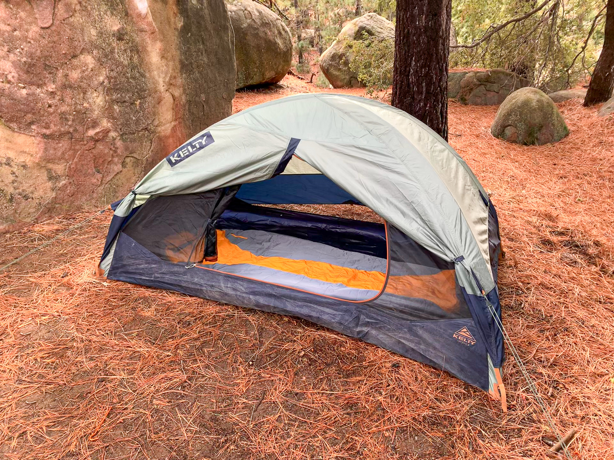 View of the Kelty Late Start 2 with the rainfly and interior door rolled back showing a sleeping bag inside. Big boulders and pine trees surround the area.