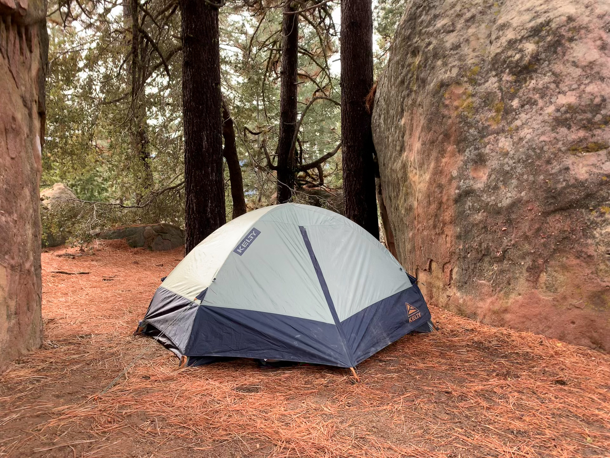 Three-quarter view of the Kelty Late Start 2 with rainfly fully zipped up. Tent is perched between to large boulders with forest pines in the background.