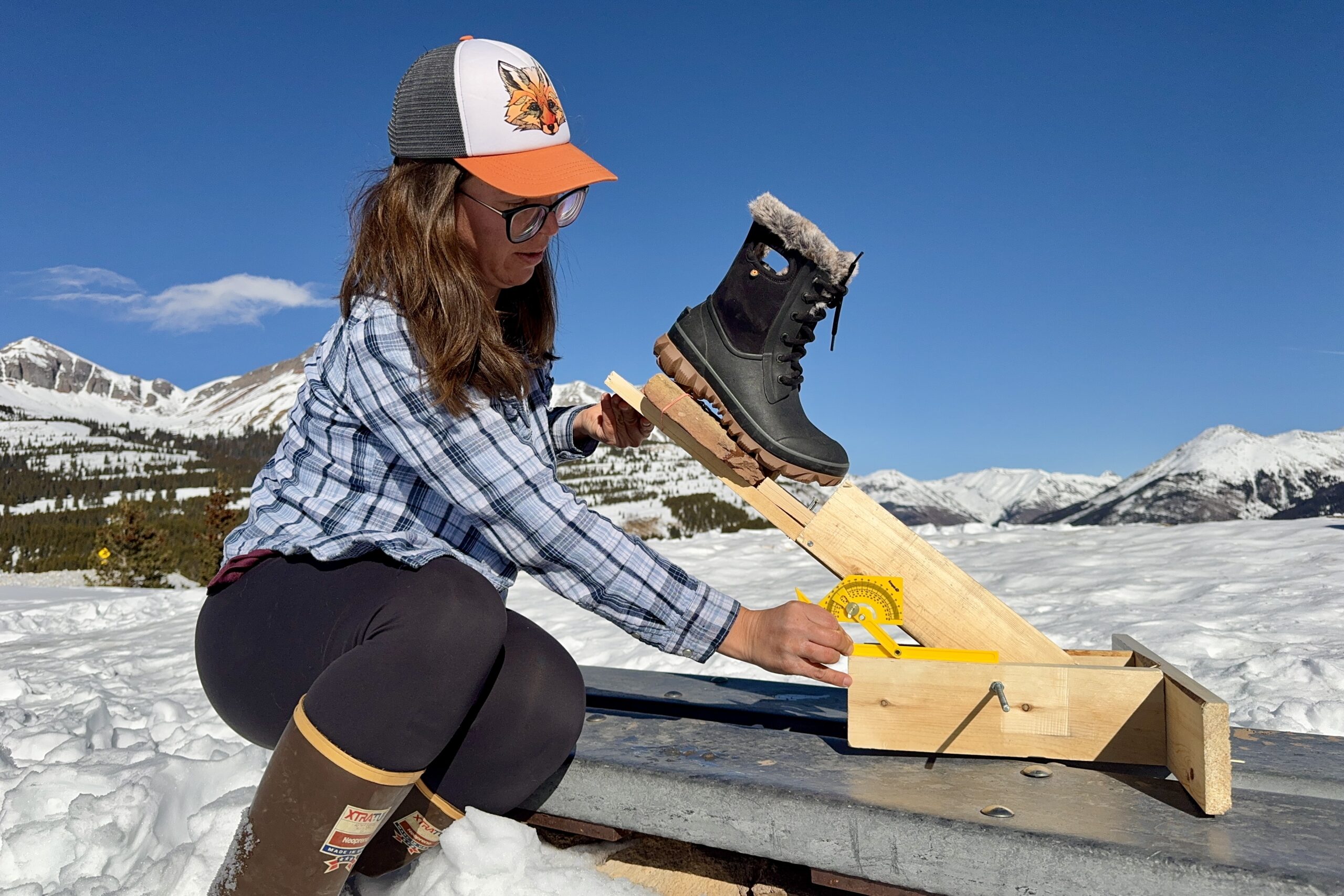 A person is measuring the traction of the Bogs Arcata boot. The boot is sitting on a rock that is perched on a wooden lever arm and the person is holding a protractor up to the device. The measurement is taking place in the snow in the mountain.