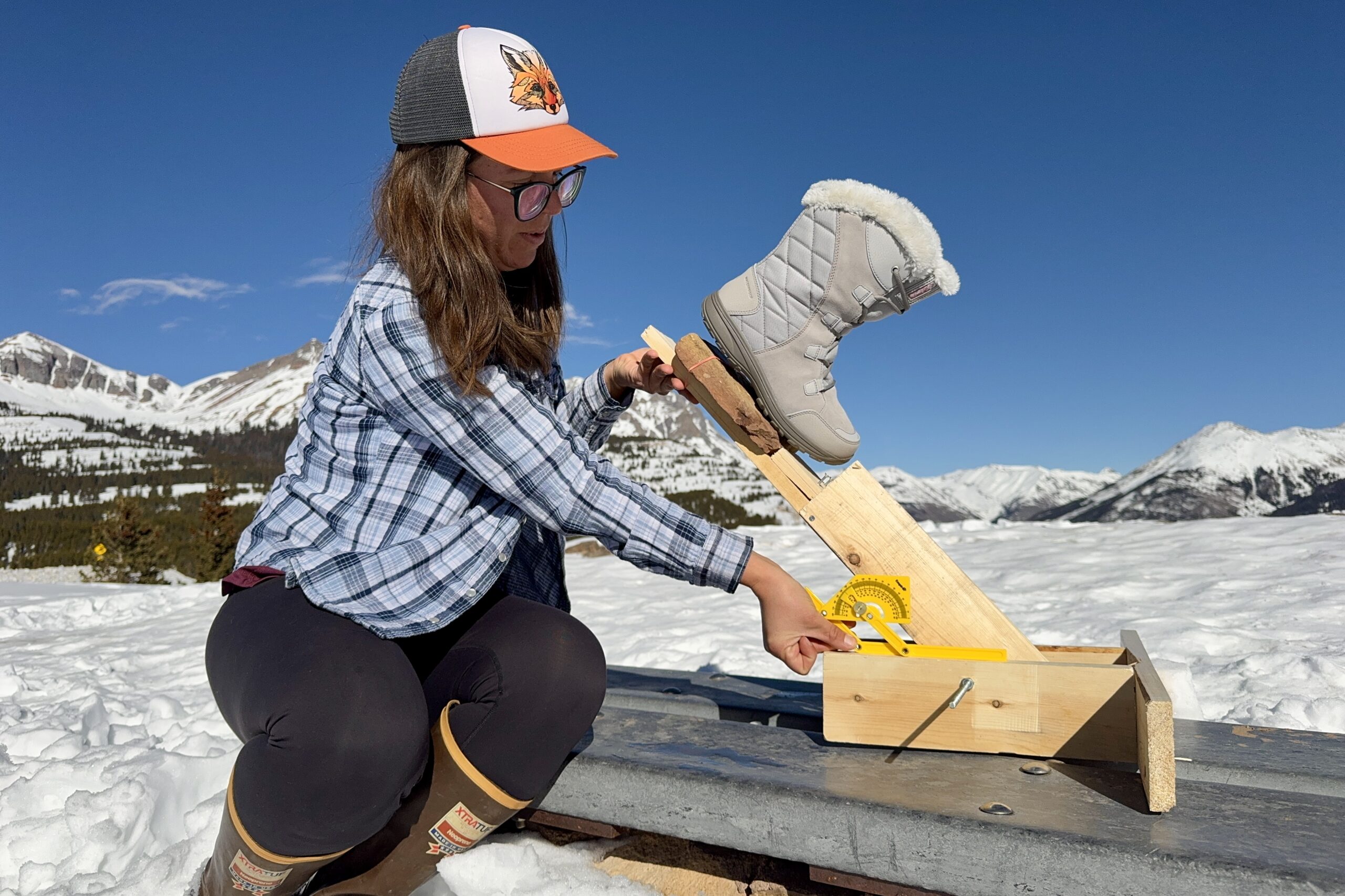 A person is measuring the traction of the Ice Maiden boot. The boot is sitting on a rock that is perched on a wooden lever arm and the person is holding a protractor up to the device. The measurement is taking place in the snow in the mountain.