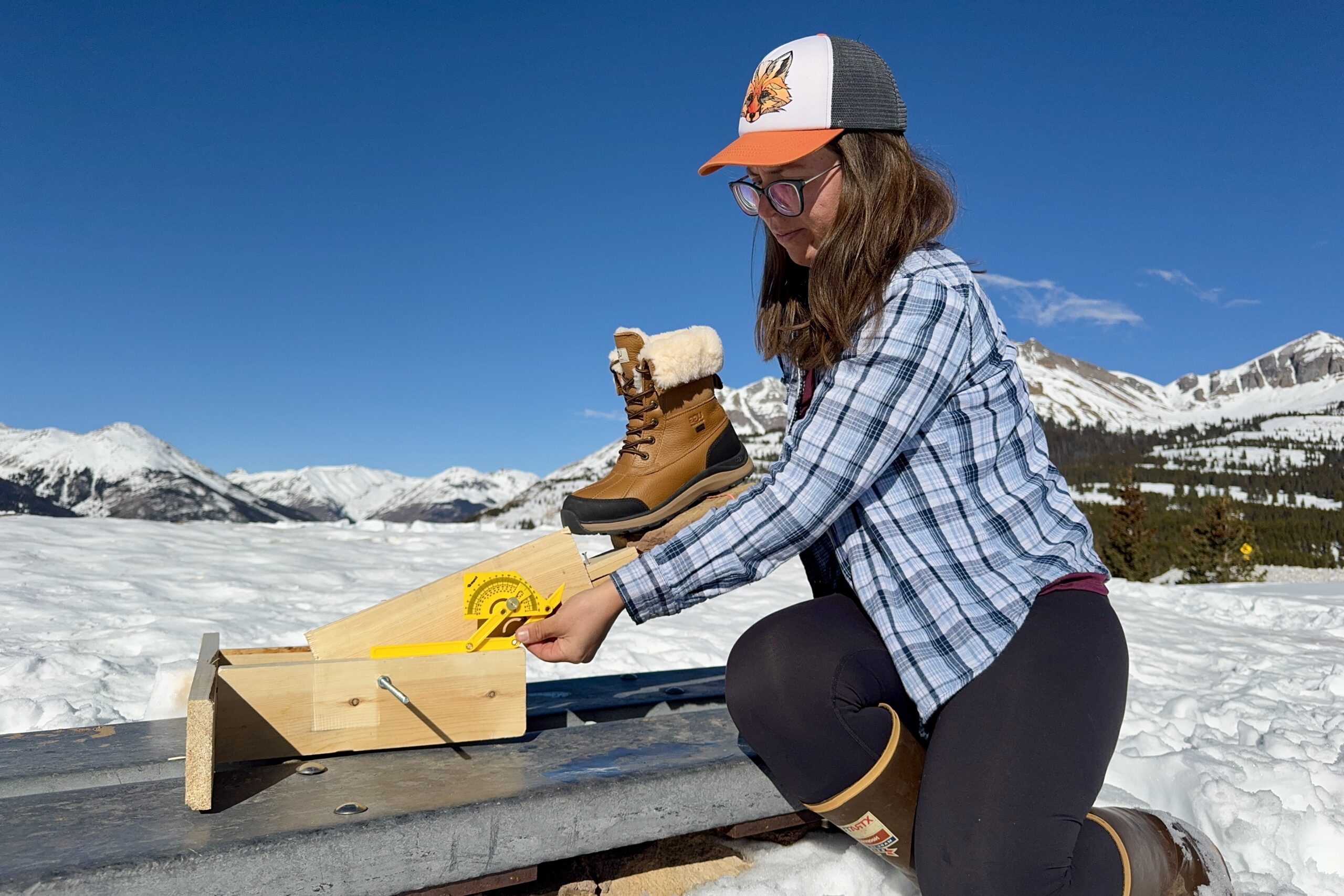 A person is measuring the traction of the UGG boot. The boot is sitting on a rock that is perched on a wooden lever arm and the person is holding a protractor up to the device. The measurement is taking place in the snow in the mountain.