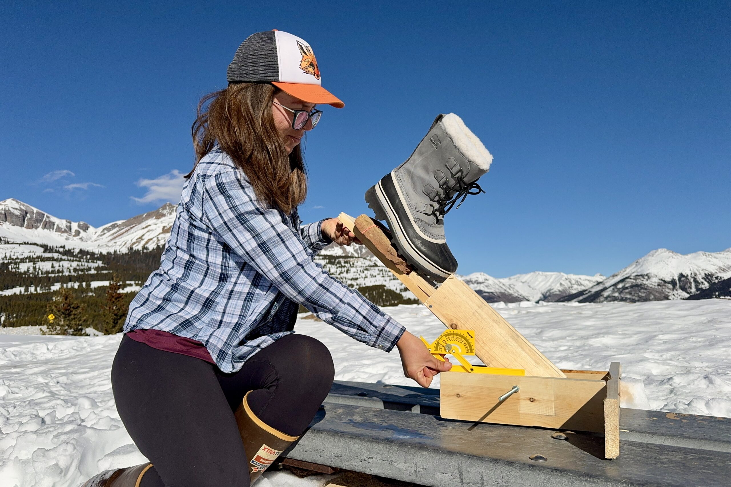 A person is measuring the traction of the Sorel boot. The boot is sitting on a rock that is perched on a wooden lever arm and the person is holding a protractor up to the device. The measurement is taking place in the snow in the mountain.