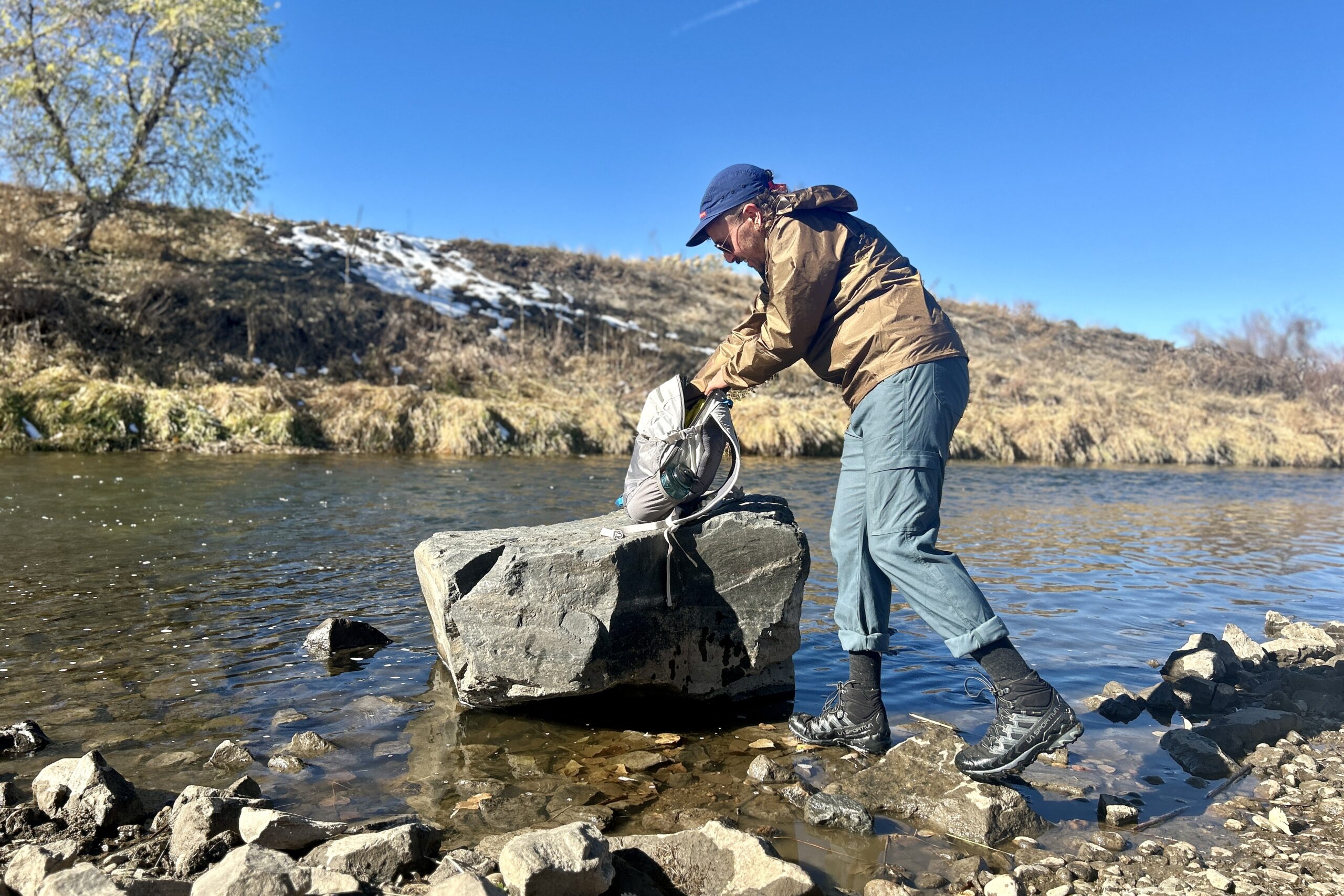 A man digs through a backpack resting on a builder at a river's edge.