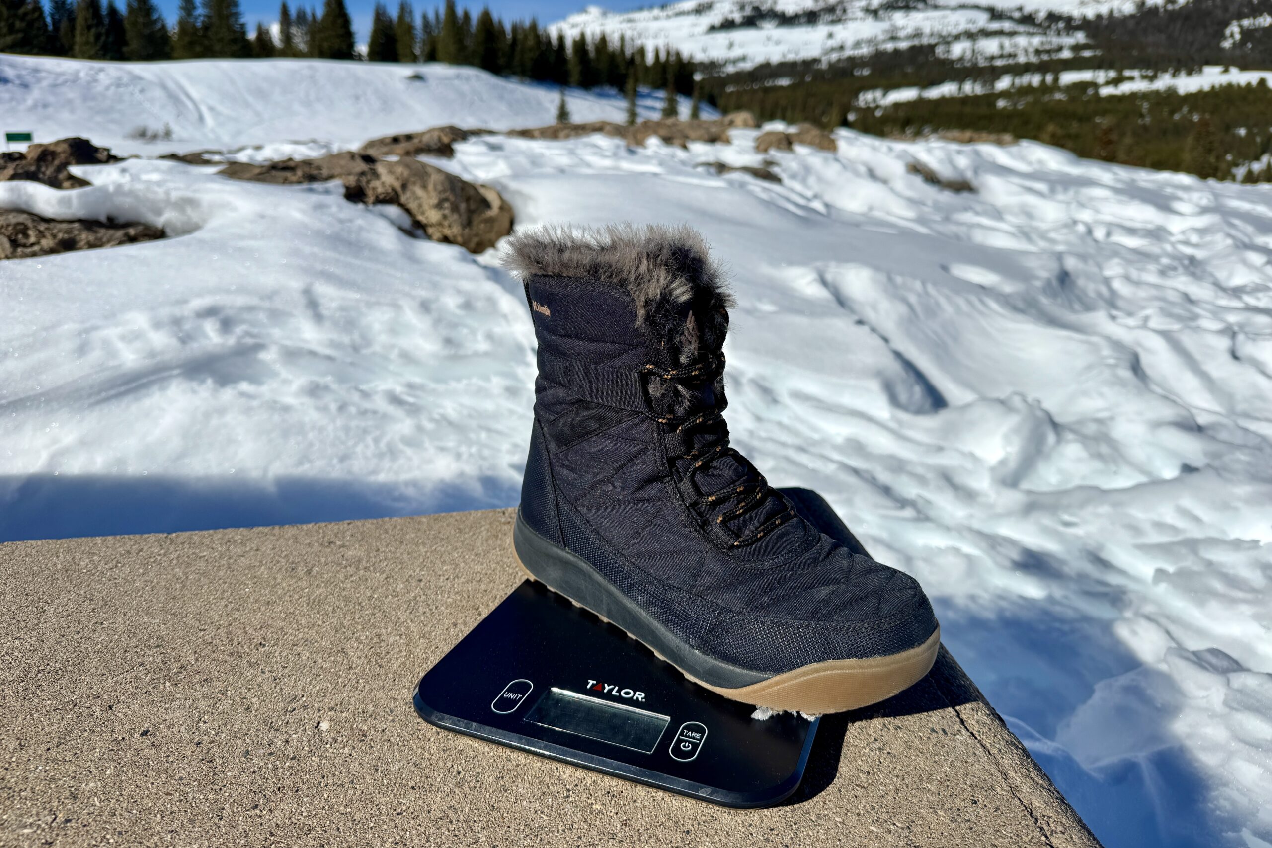 A close up of the Minx Shorty boot sitting on a kitchen scale. The scale is sitting on a concrete wall in the mountains and there is snow covering the ground.