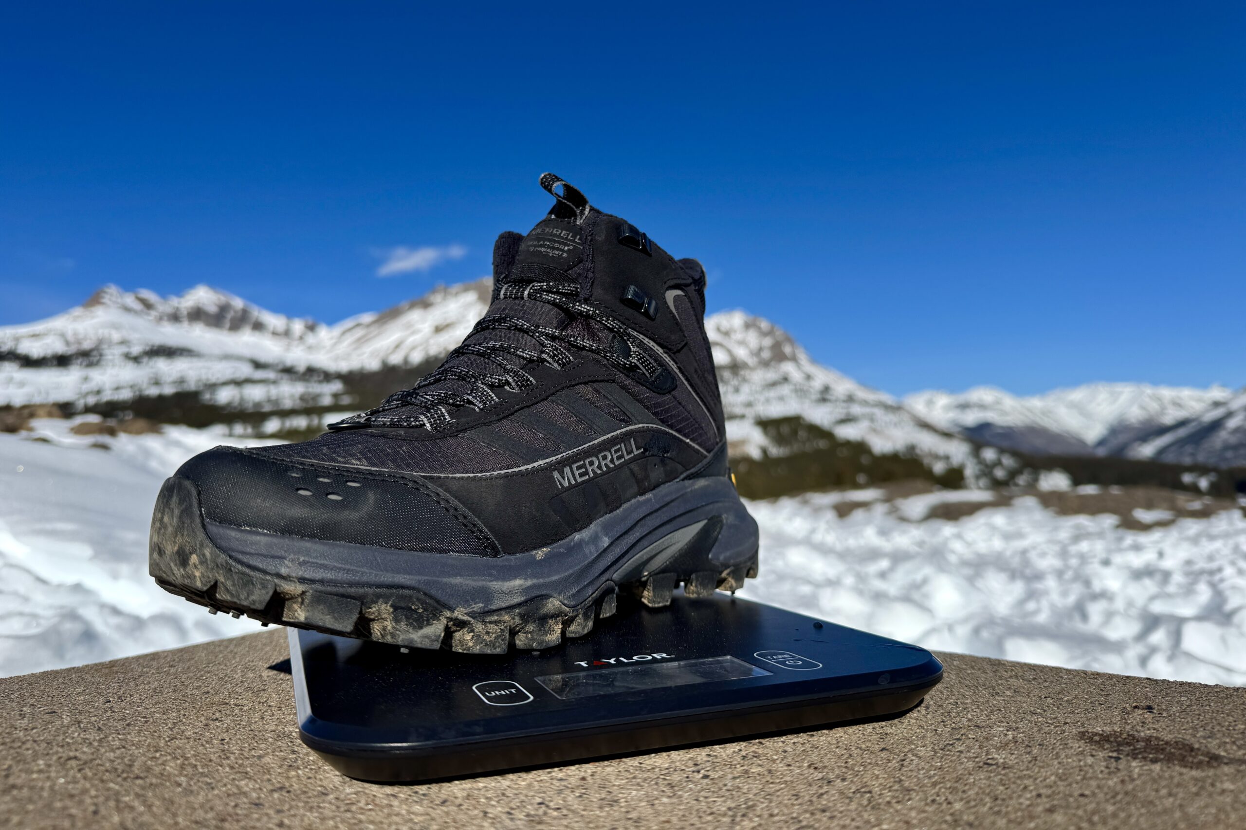 A close up of the Merrell boot sitting on a kitchen scale. The scale is sitting on a concrete wall in the mountains and there is snow covering the ground.