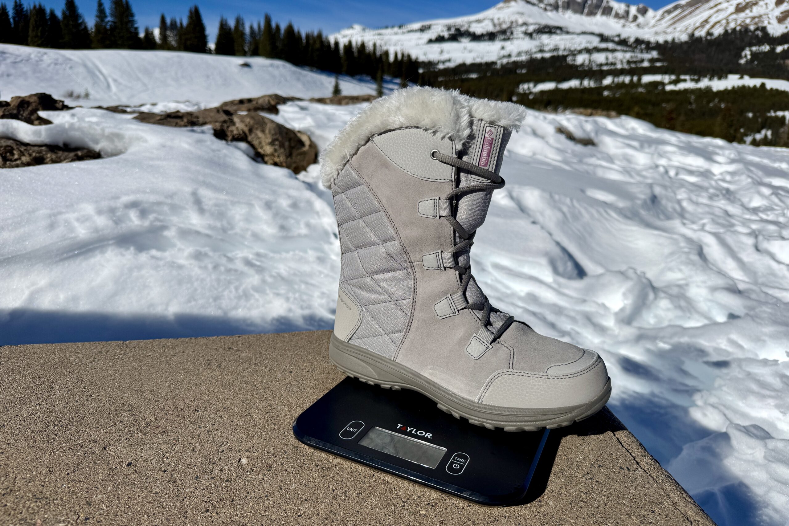 A close up of the Ice Maiden boot sitting on a kitchen scale. The scale is sitting on a concrete wall in the mountains and there is snow covering the ground.