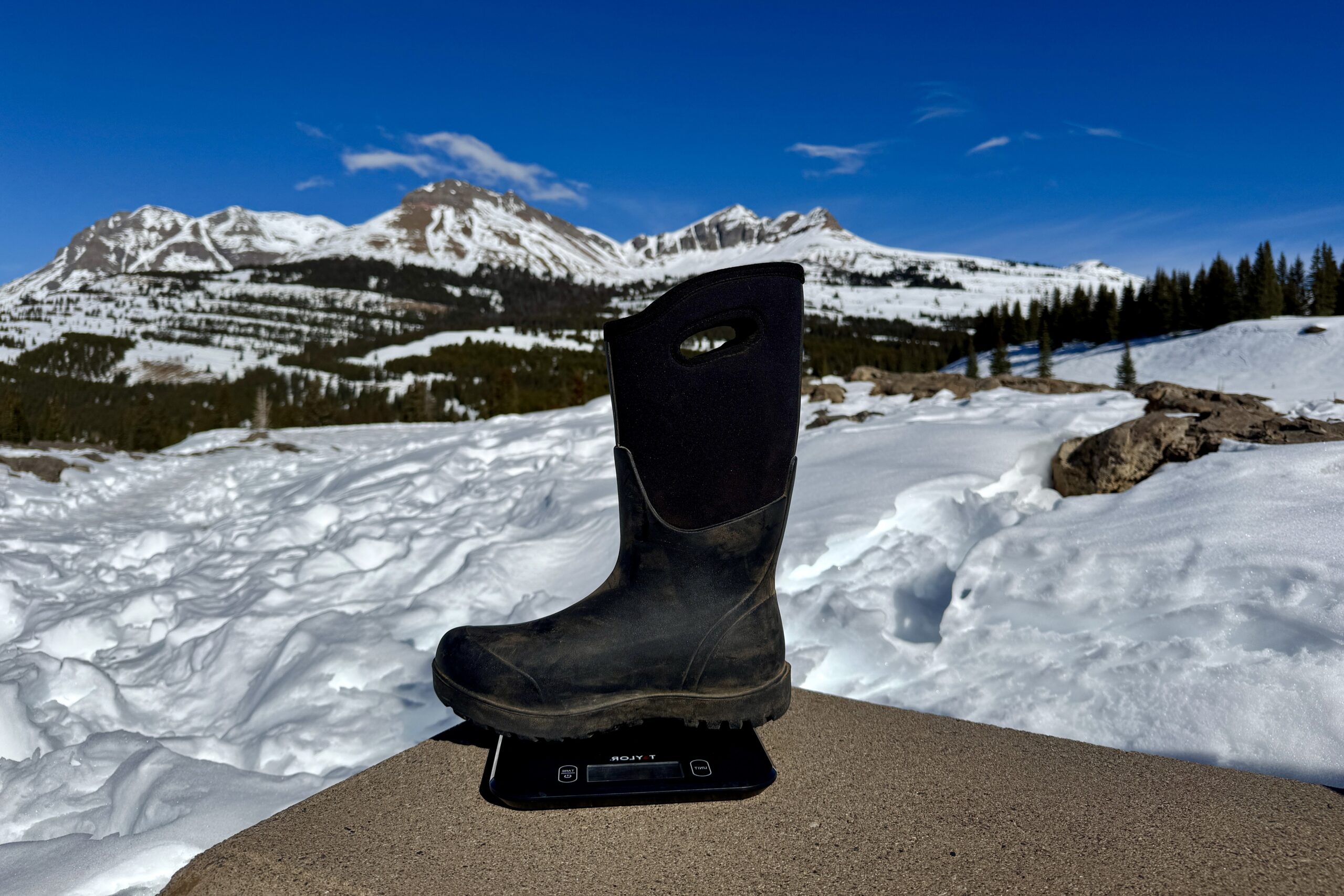 A close up of the Bogs Neo Classic Tall boot sitting on a kitchen scale. The scale is sitting on a concrete wall in the mountains and there is snow covering the ground.