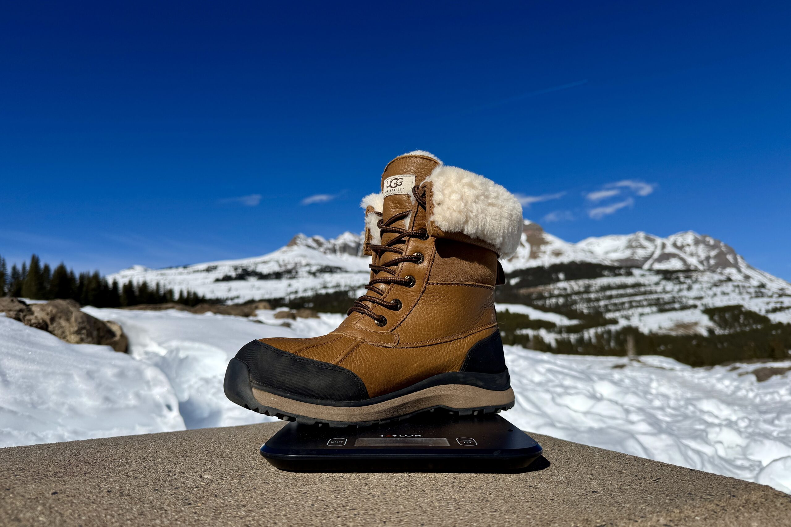 A close up of the UGG boot sitting on a kitchen scale. The scale is sitting on a concrete wall in the mountains and there is snow covering the ground.