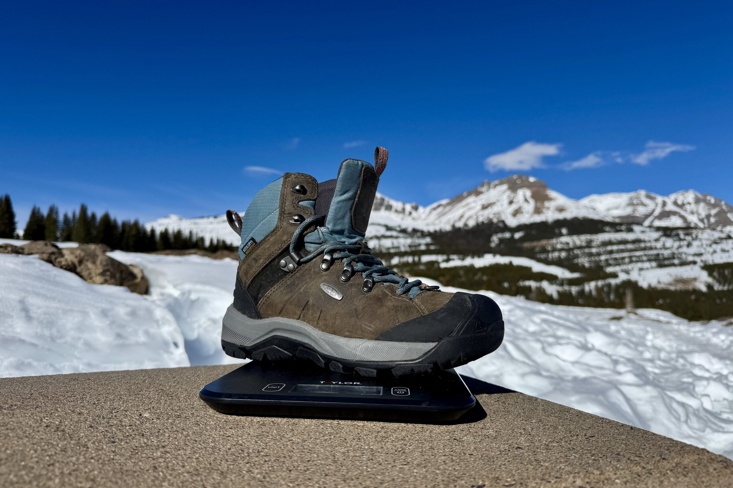 A close up of the Keen boot sitting on a kitchen scale. The scale is sitting on a concrete wall in the mountains and there is snow covering the ground.