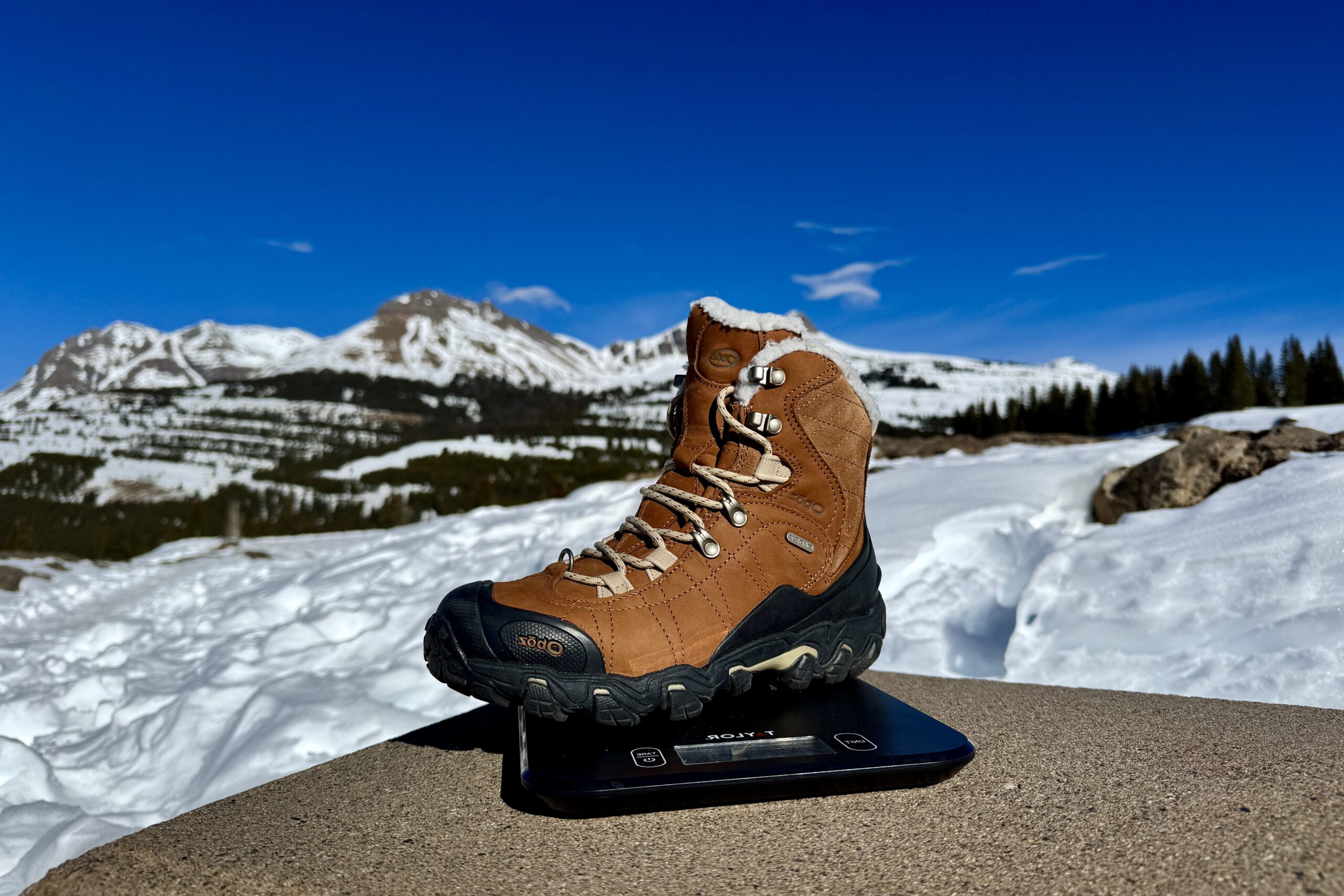 A close up of the Oboz boot sitting on a kitchen scale. The scale is sitting on a concrete wall in the mountains and there is snow covering the ground.