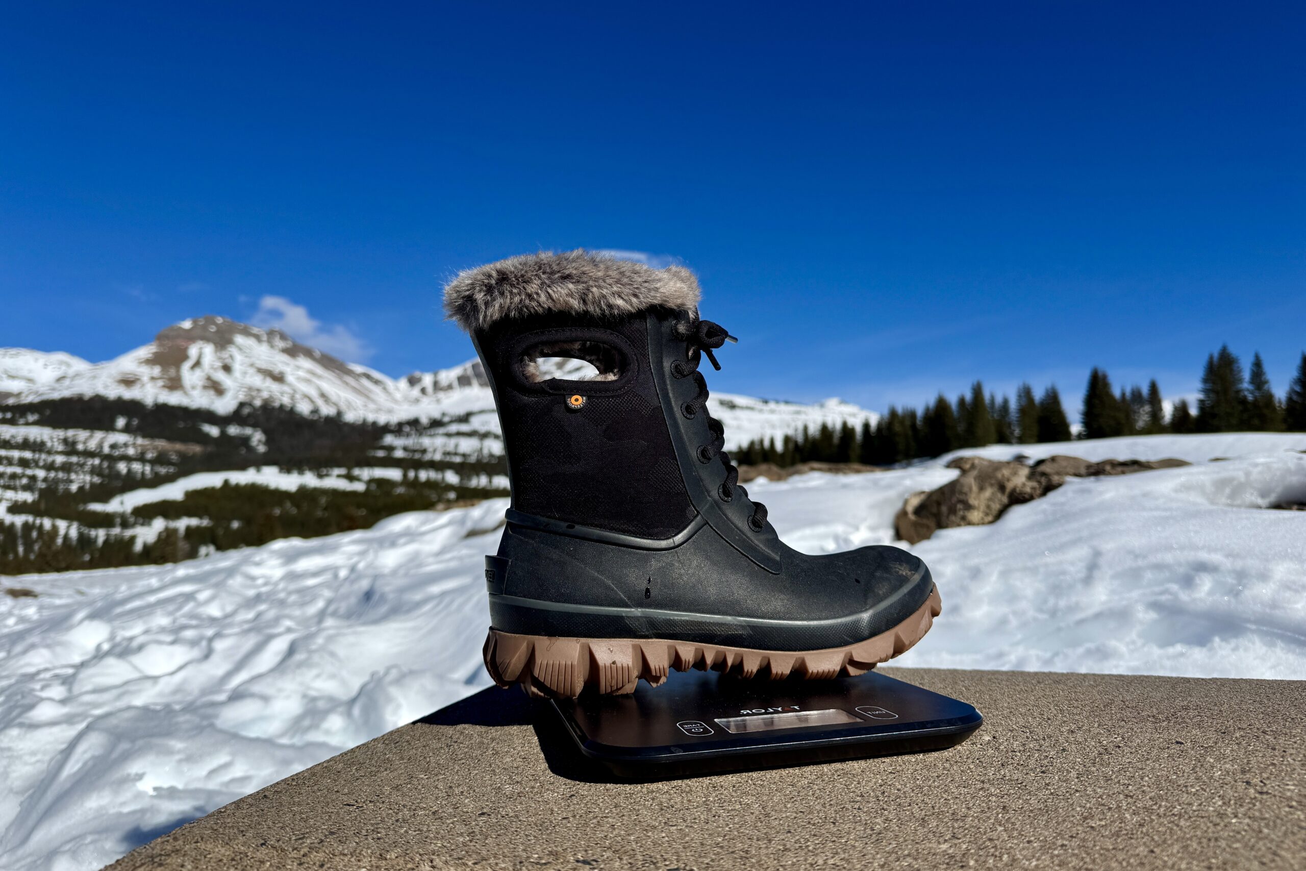 A close up of the Bogs Arcata boot sitting on a kitchen scale. The scale is sitting on a concrete wall in the mountains and there is snow covering the ground.