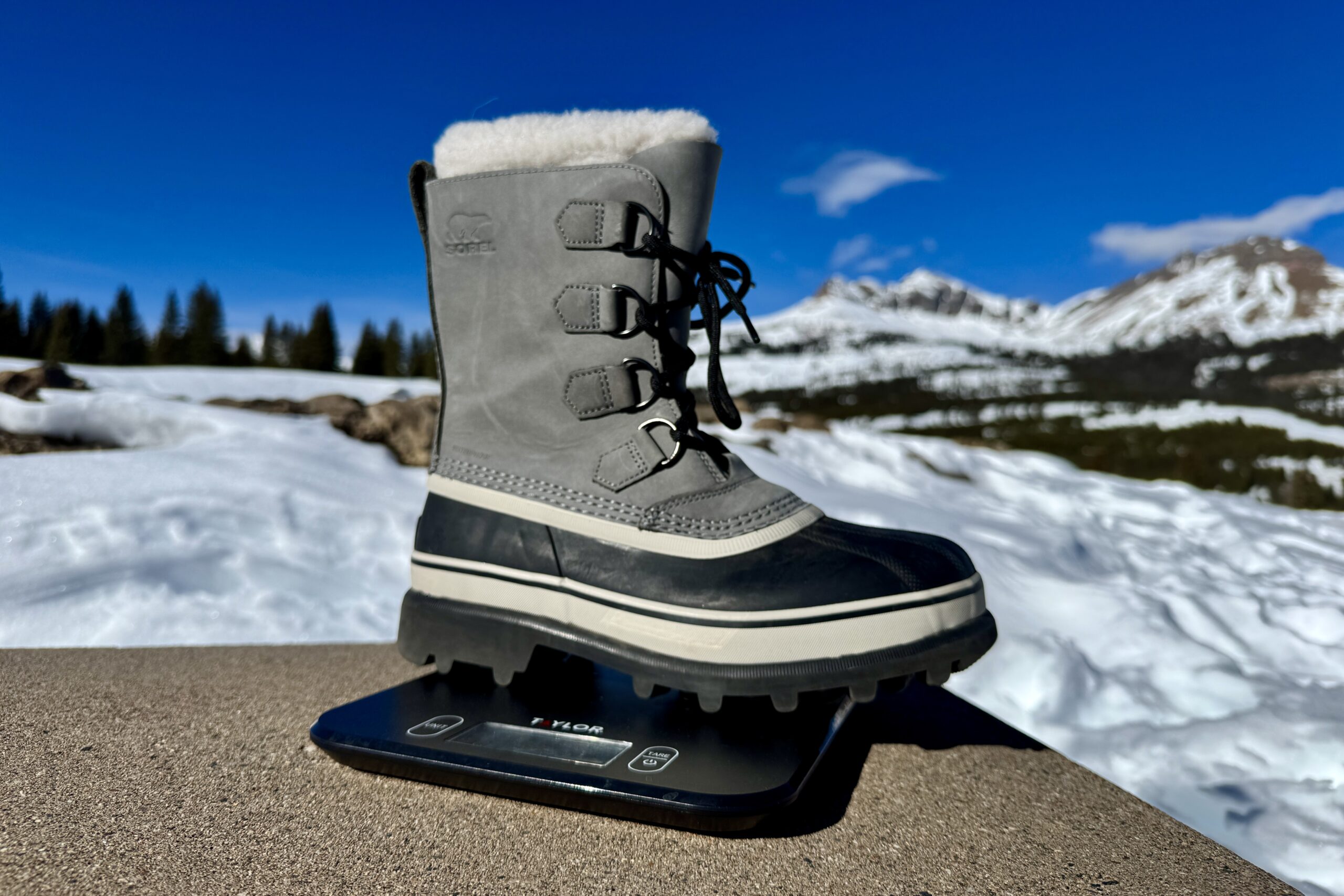 A close up of the Sorel boot sitting on a kitchen scale. The scale is sitting on a concrete wall in the mountains and there is snow covering the ground.