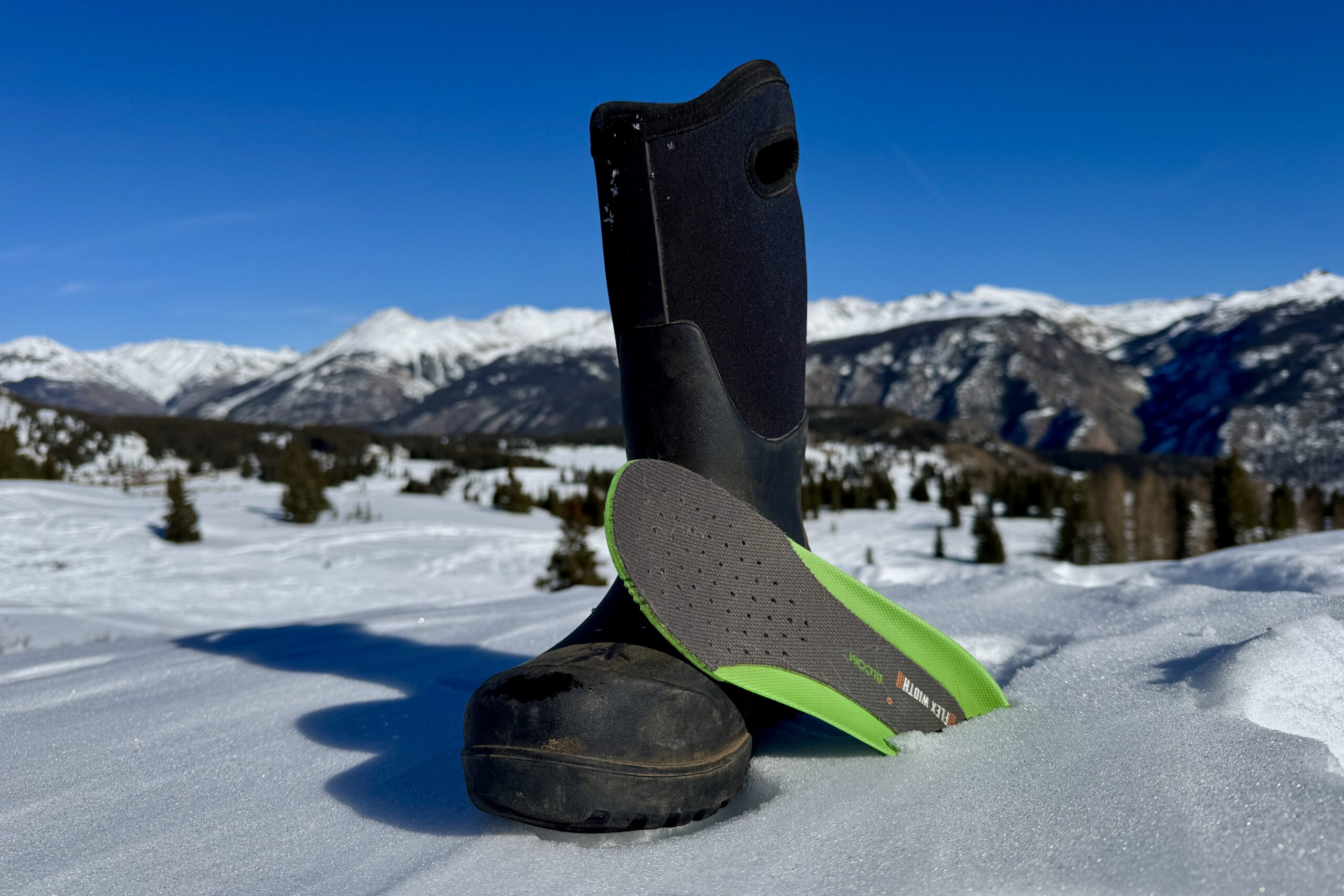 A close up of the Bogs Neo Classic Tall boot with the insole sitting next to it. They are set on some snow in the mountains.