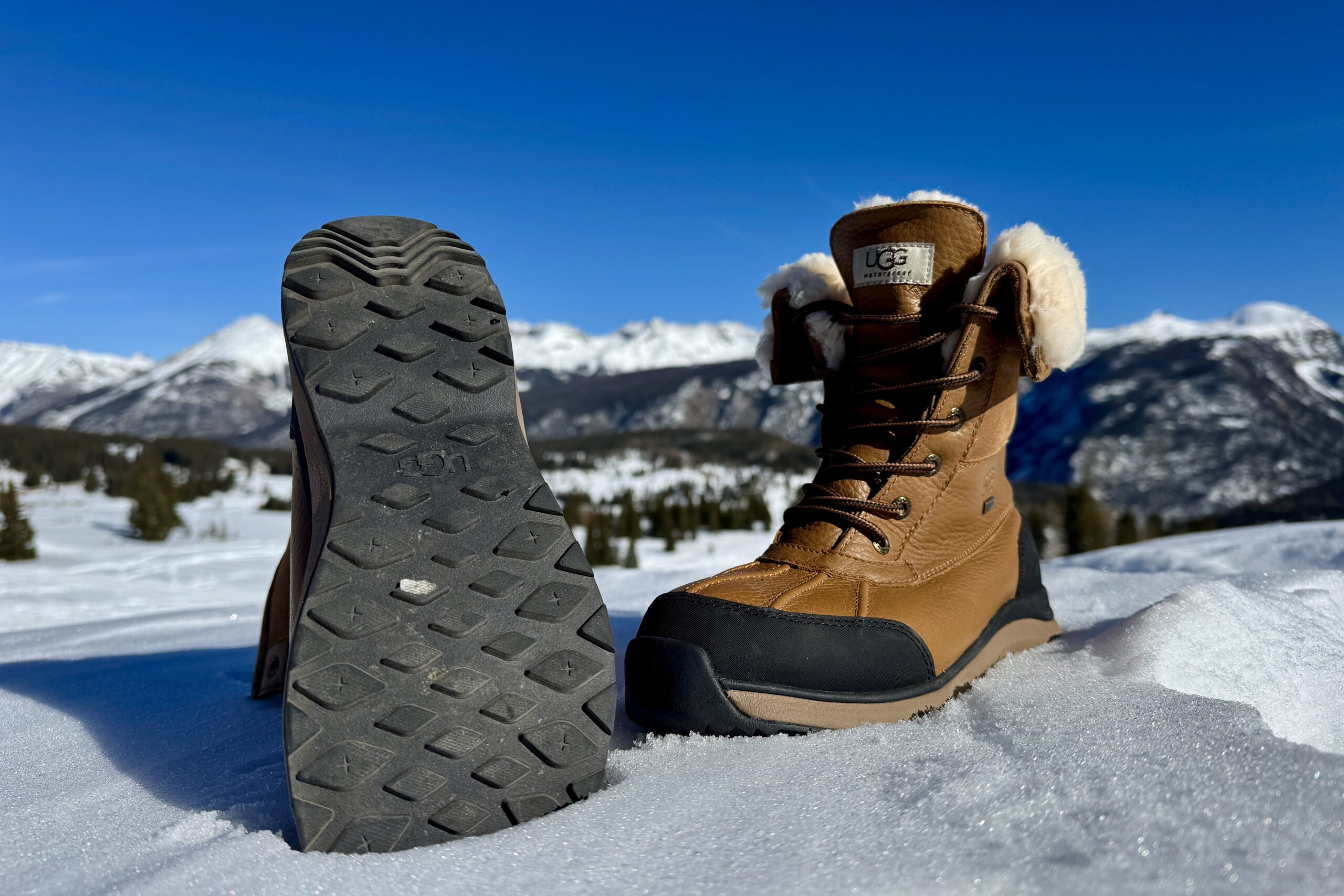 A close up of the tread on the bottom of the UGG boot. It is sitting in the snow in the mountains.