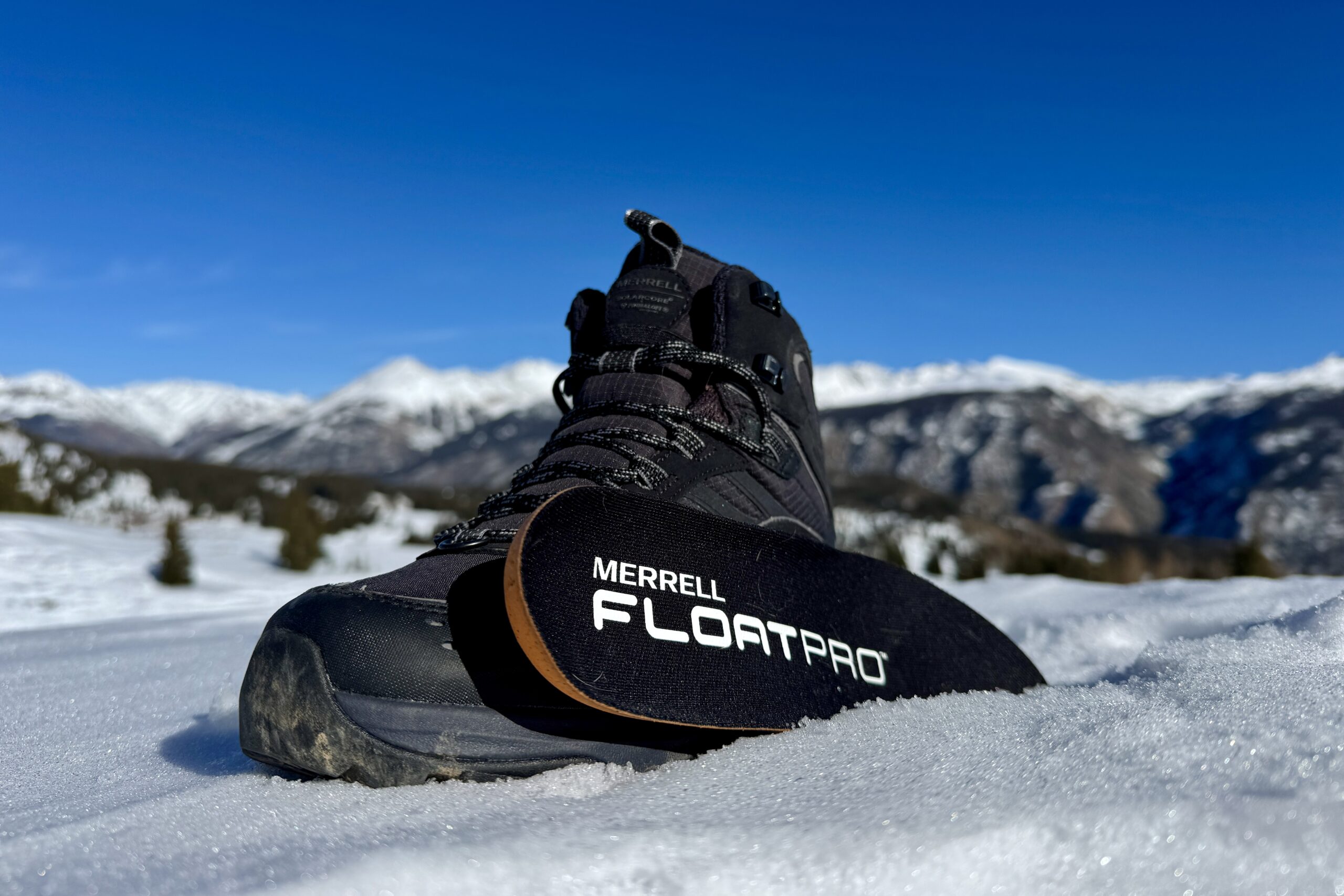 A close up of the Merrell boot with the insole sitting next to it. They are set on some snow in the mountains.