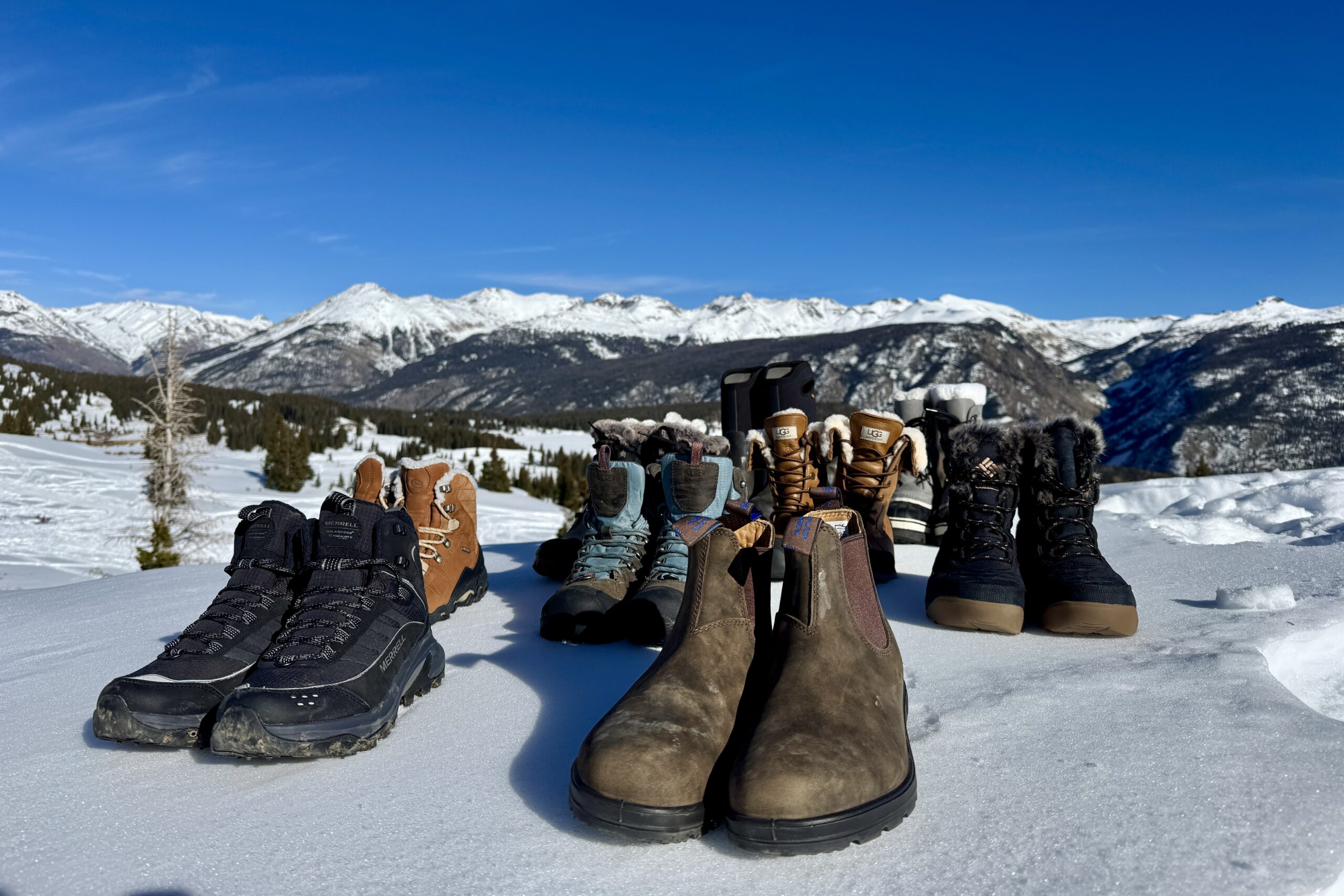 All the boots we tested are lined up on top of the snow with mountains in the background.