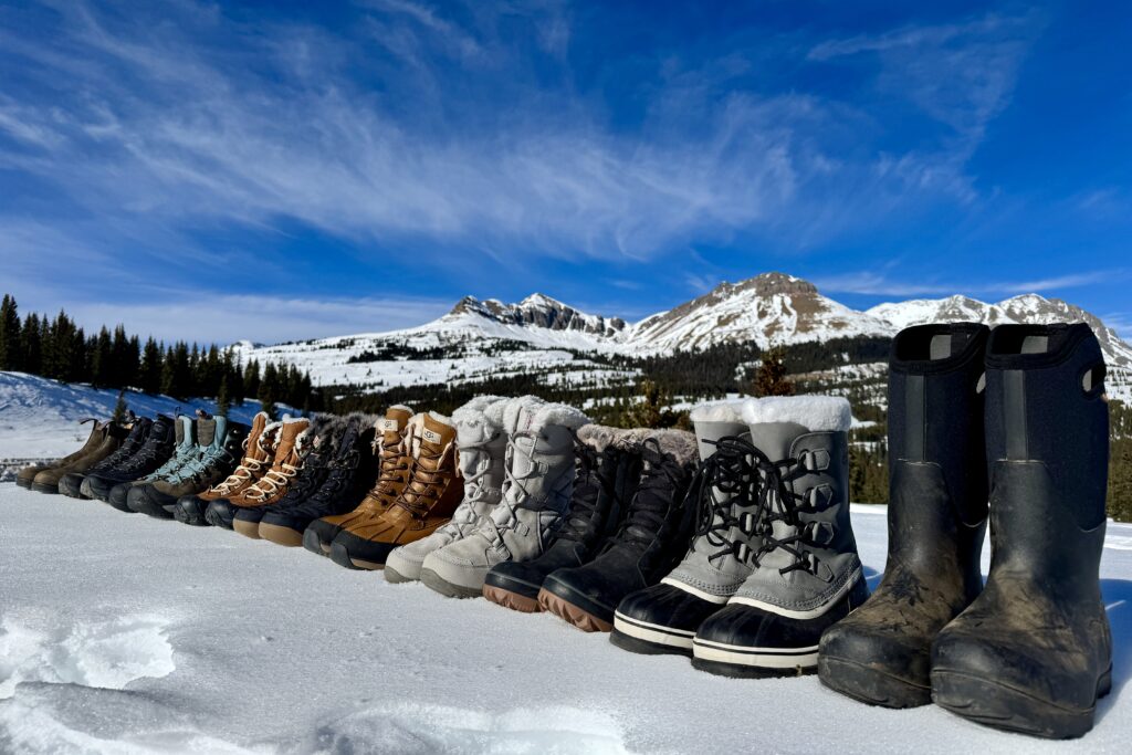 All the boots we tested are lined up on top of the snow with mountains in the background.