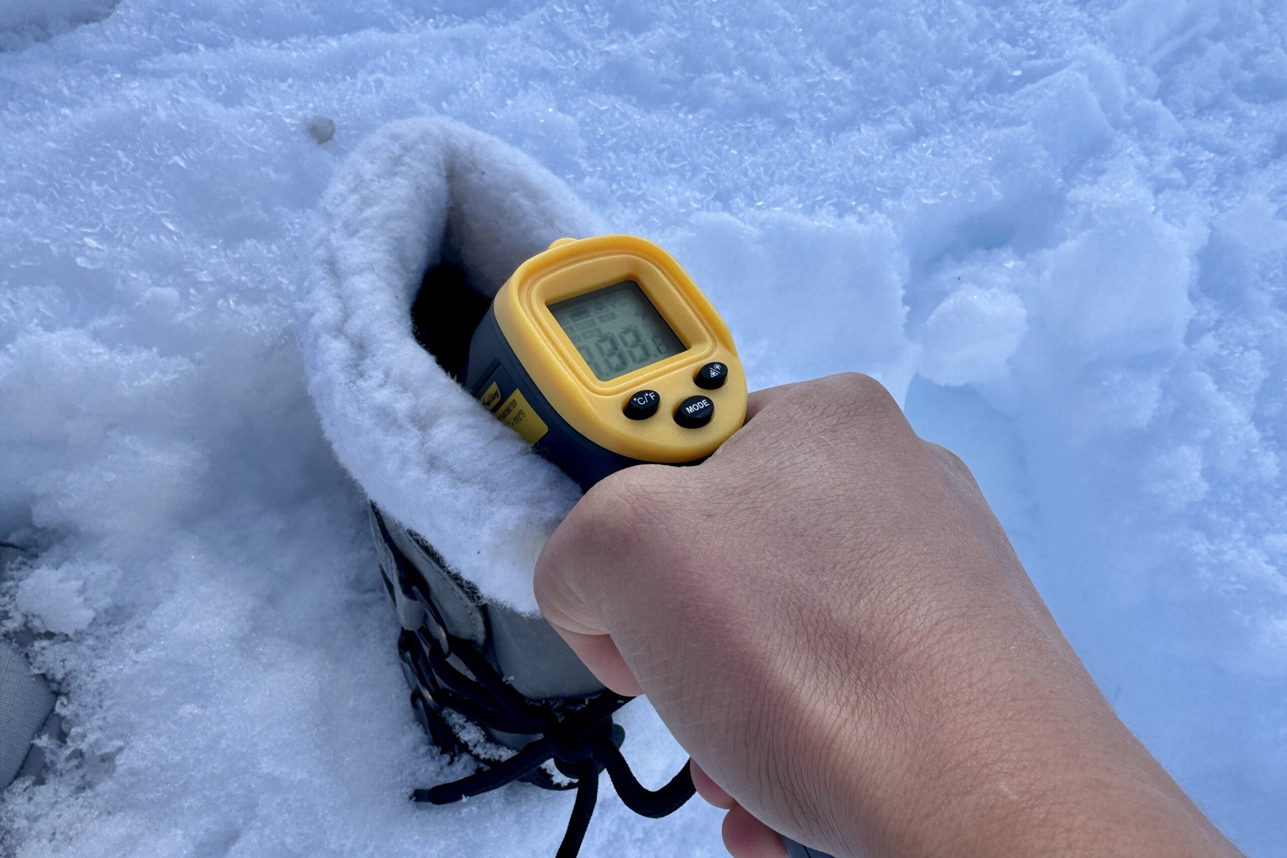 Close up of a hand pointing a laser thermometer into the Sorel boot which is sitting in the snow.