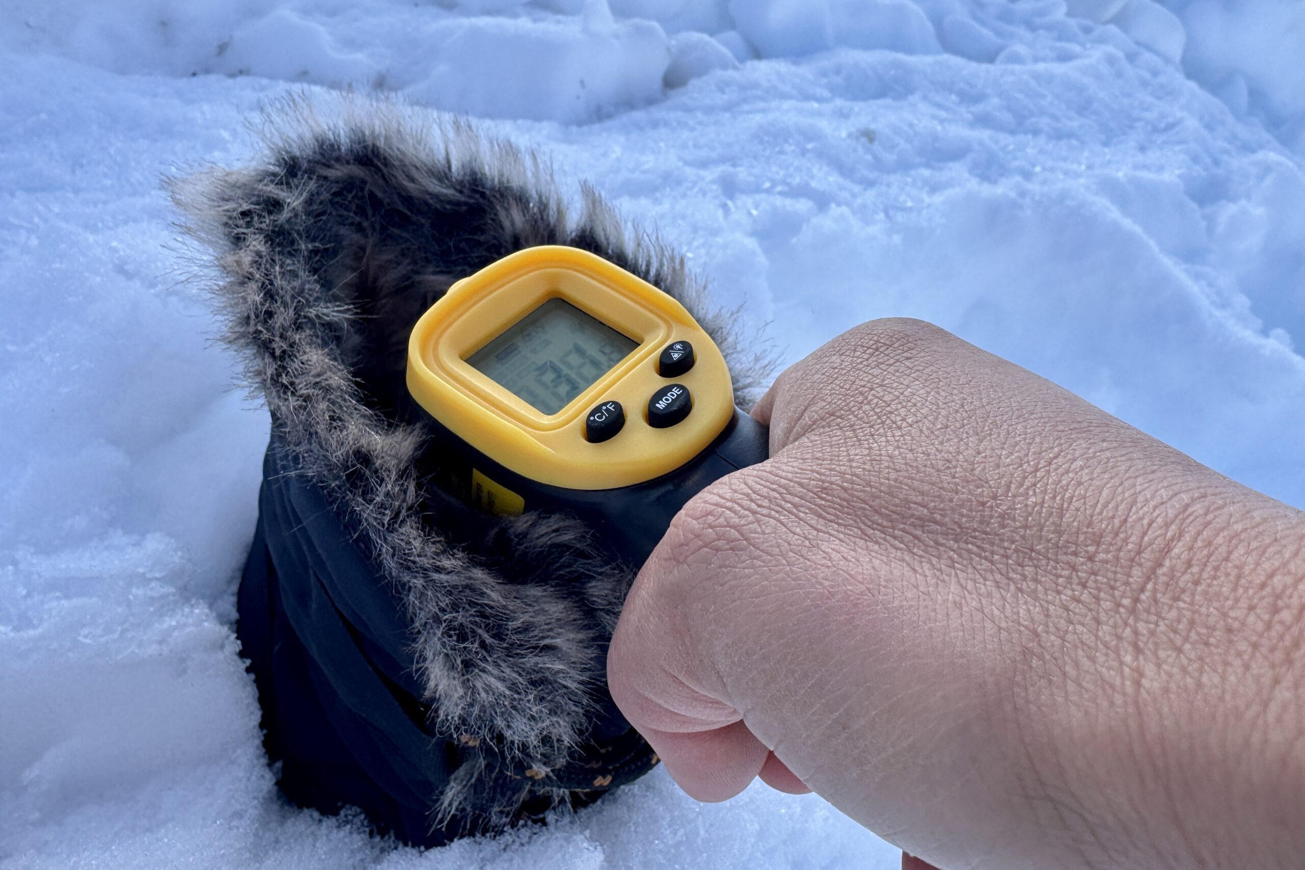 Close up of a hand pointing a laser thermometer into the Bogs Arcata boot which is sitting in the snow.