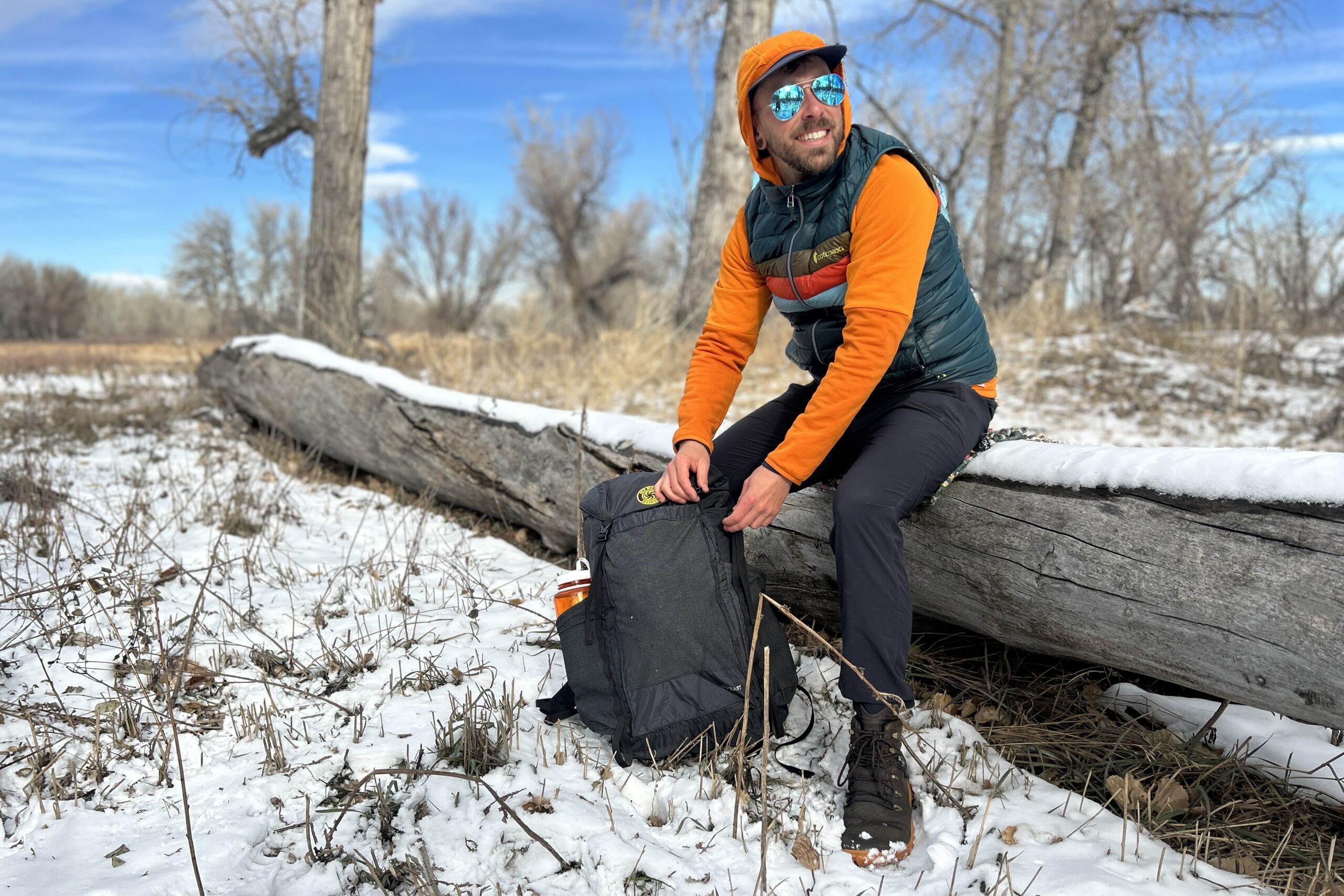 A smiling man sits on a log with his backpack on the ground in a wintry wilderness setting.