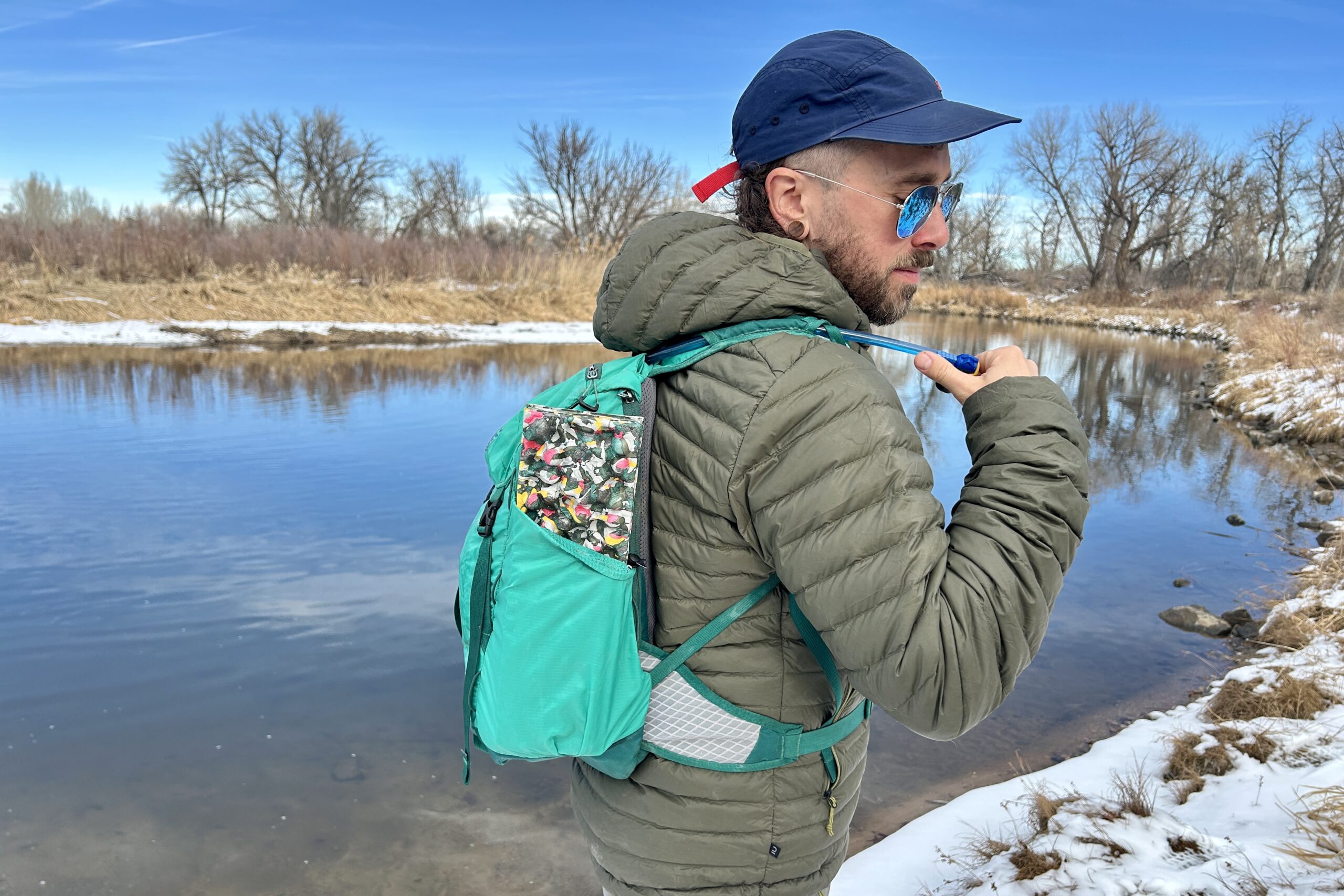 Closeup image of a man reaching for a drinking tube attached to his backpack while hiking next to a snowy river.