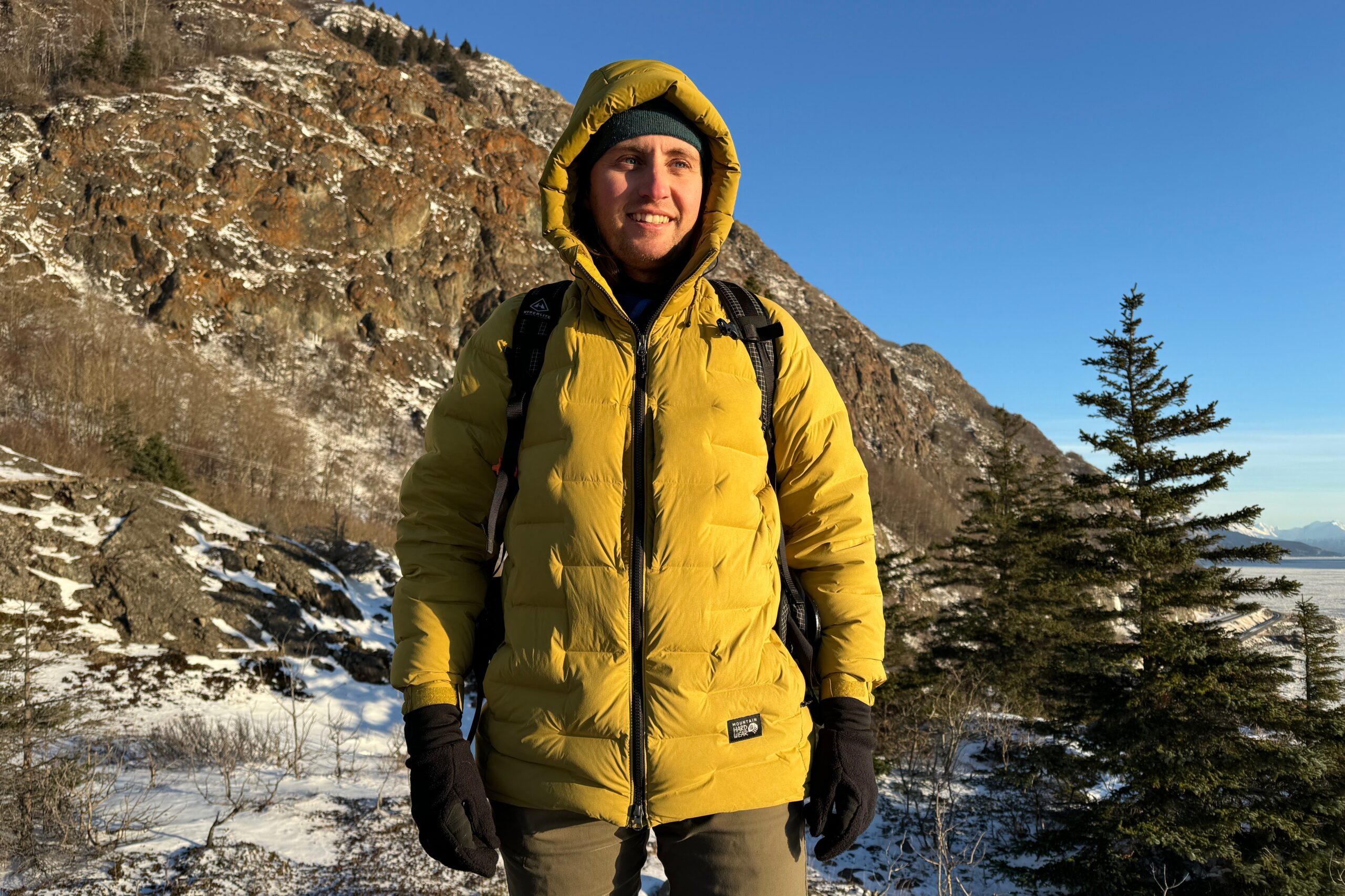 A man stands in a yellow down jacket in front of a rocky ridge.