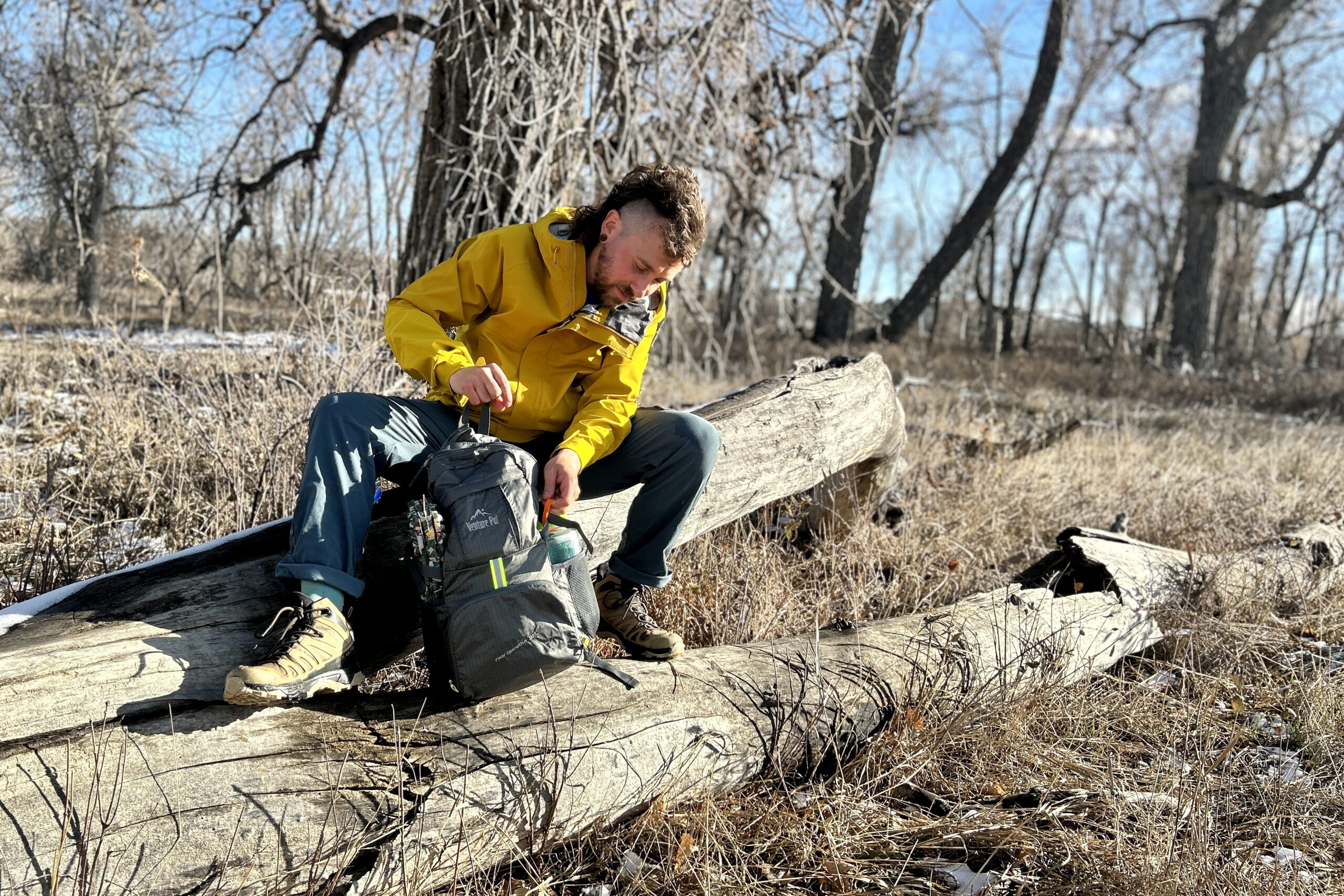 A man sits on a log and digs through a backpack in a wintry forest landscape on a sunny day.