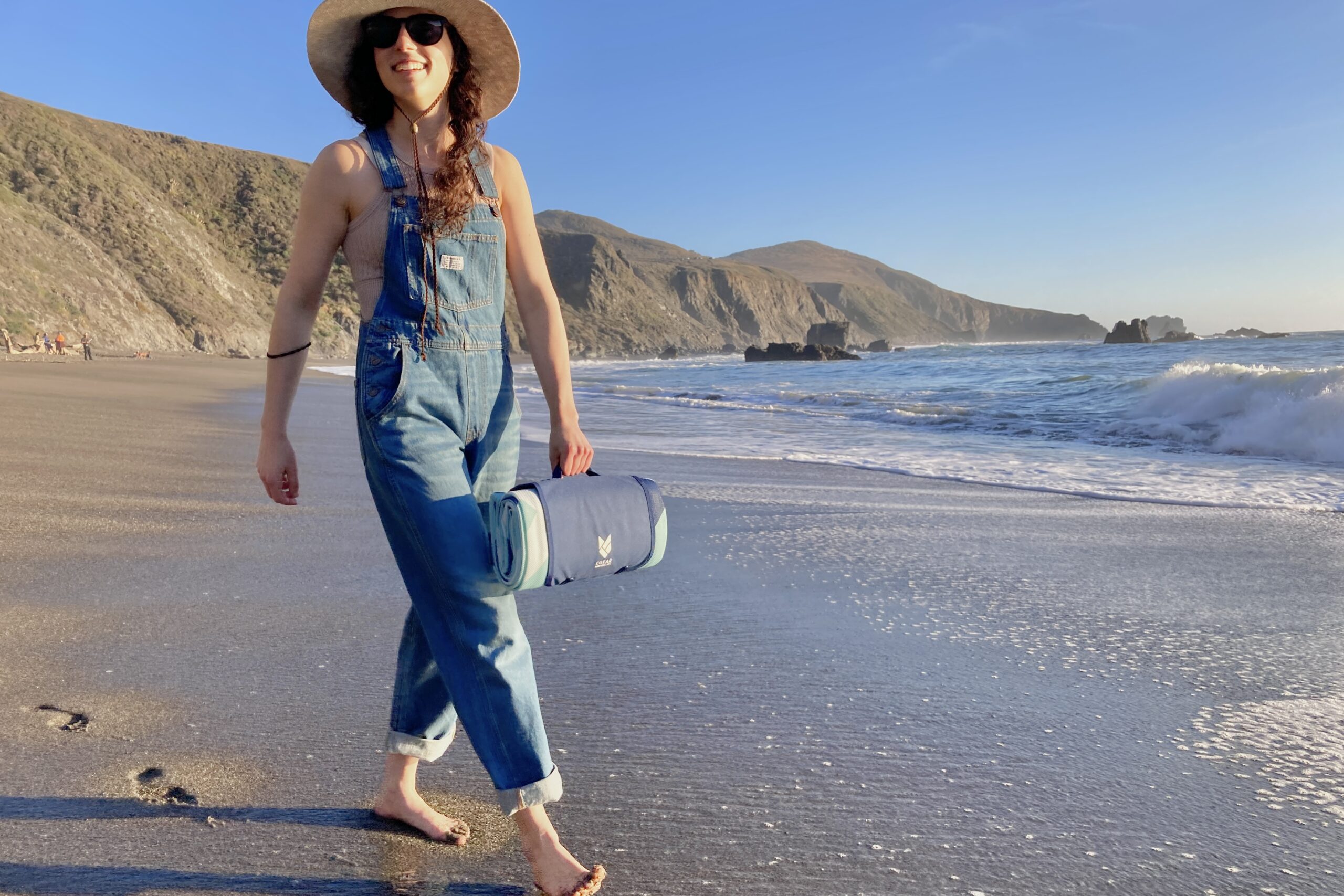 Woman walking on the beach carrying the CGear Multimats Sandlite Sand-free Mat in its handy carry case.