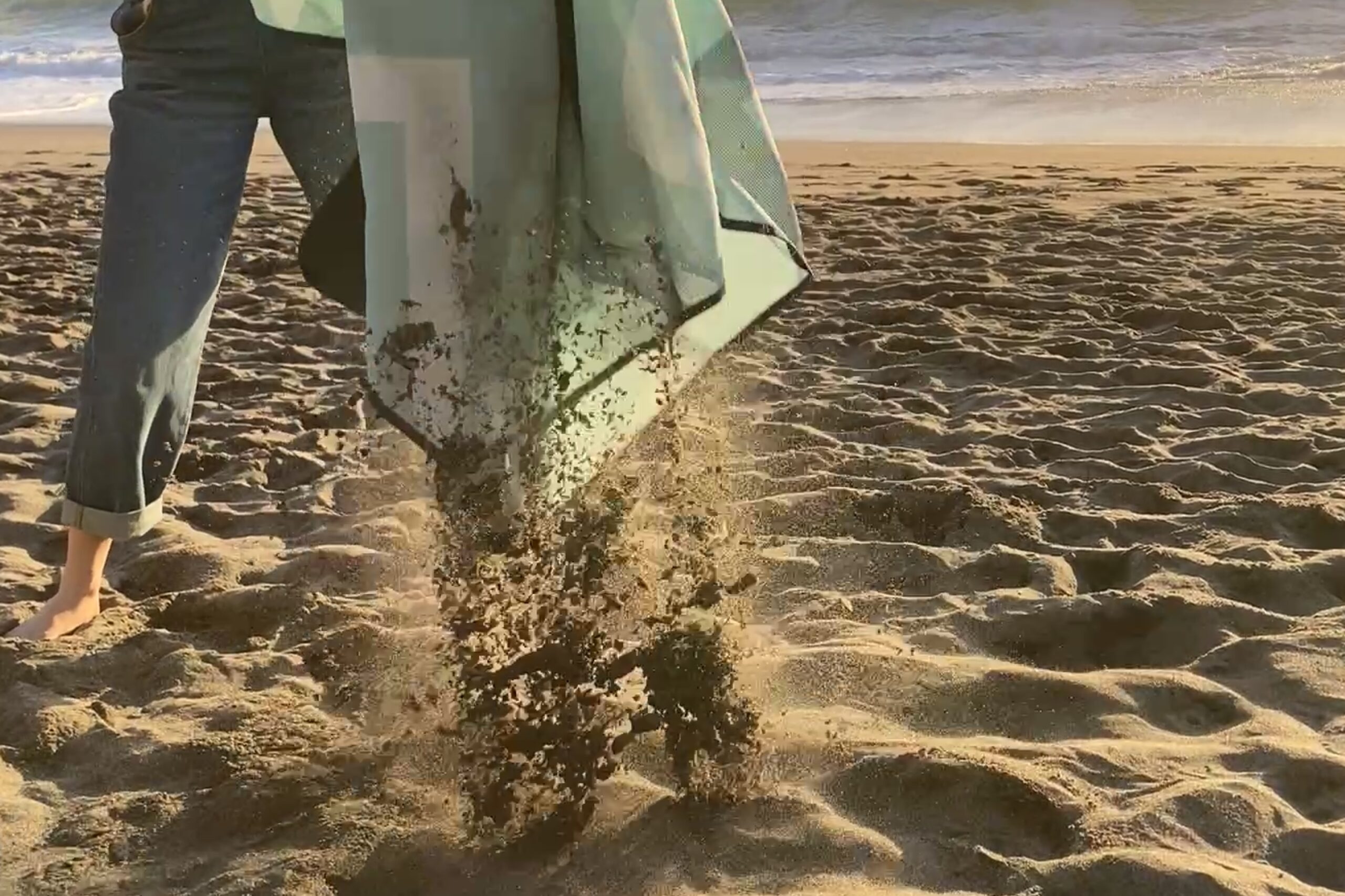 Person shaking sand off of the CGear Multimats Sandlite Sand-Free Mat at the beach.